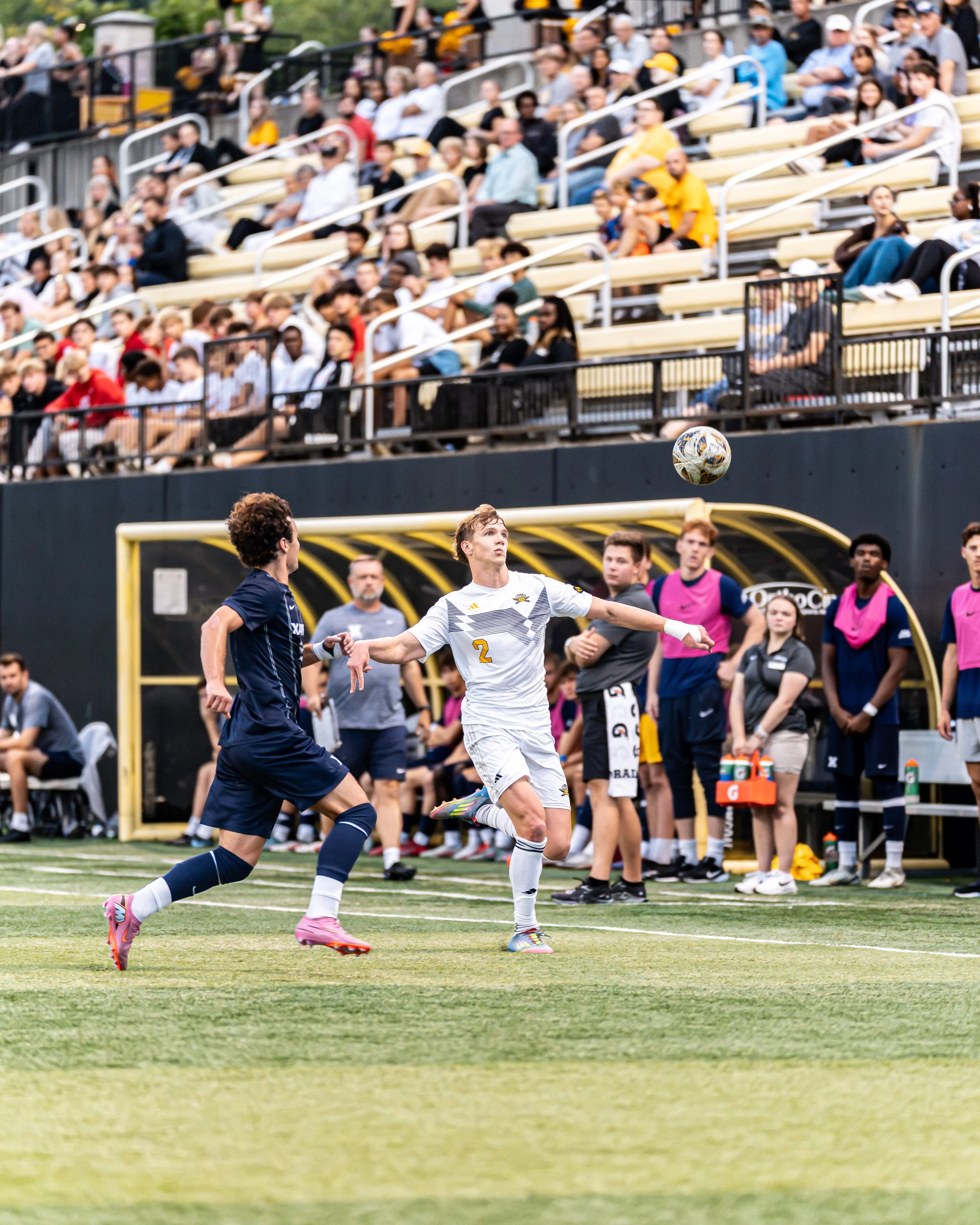 A soccer match with players on the field, one player in white jersey #2 preparing to kick the ball, and an opposing player in navy jersey nearby. Spectators are visible in the stands behind the field.