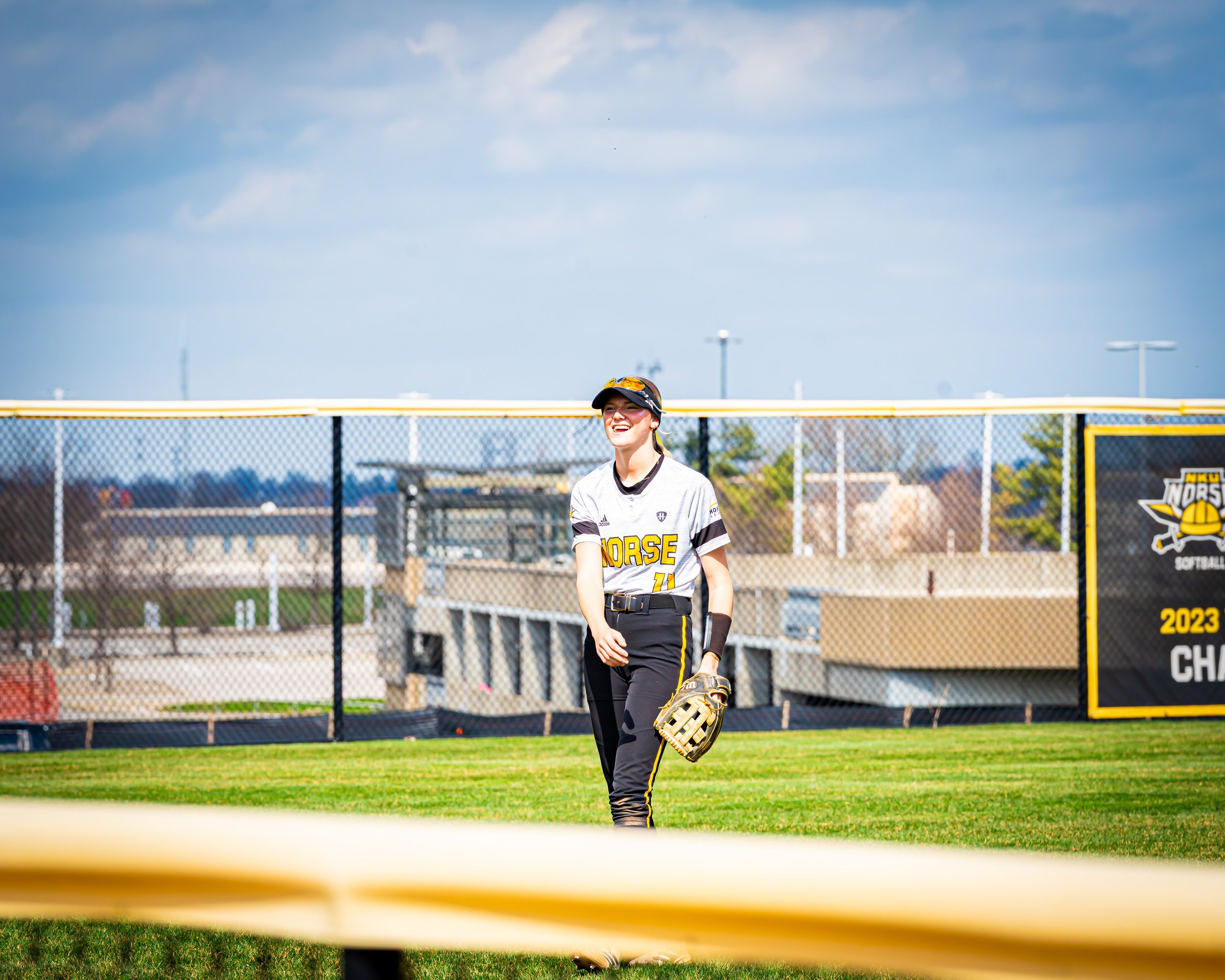 A young female softball player in a white jersey with yellow and black details, holding a glove, walking on a softball field during daytime with an overcast sky.