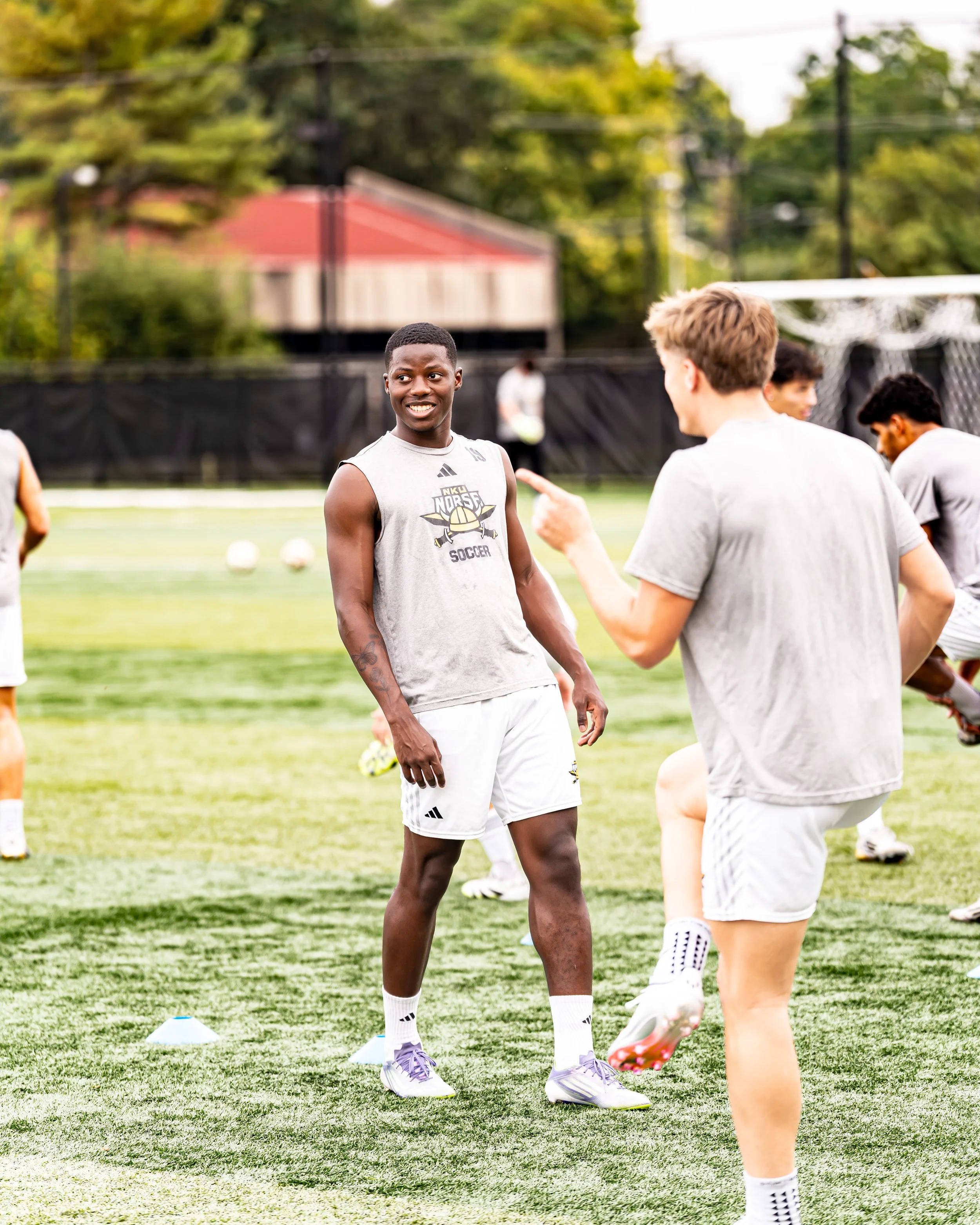 Soccer players practicing on a field with green grass, cones, and a goalpost in the background. One player smiling and wearing a sleeveless shirt, white shorts, and cleats, talking to another player.