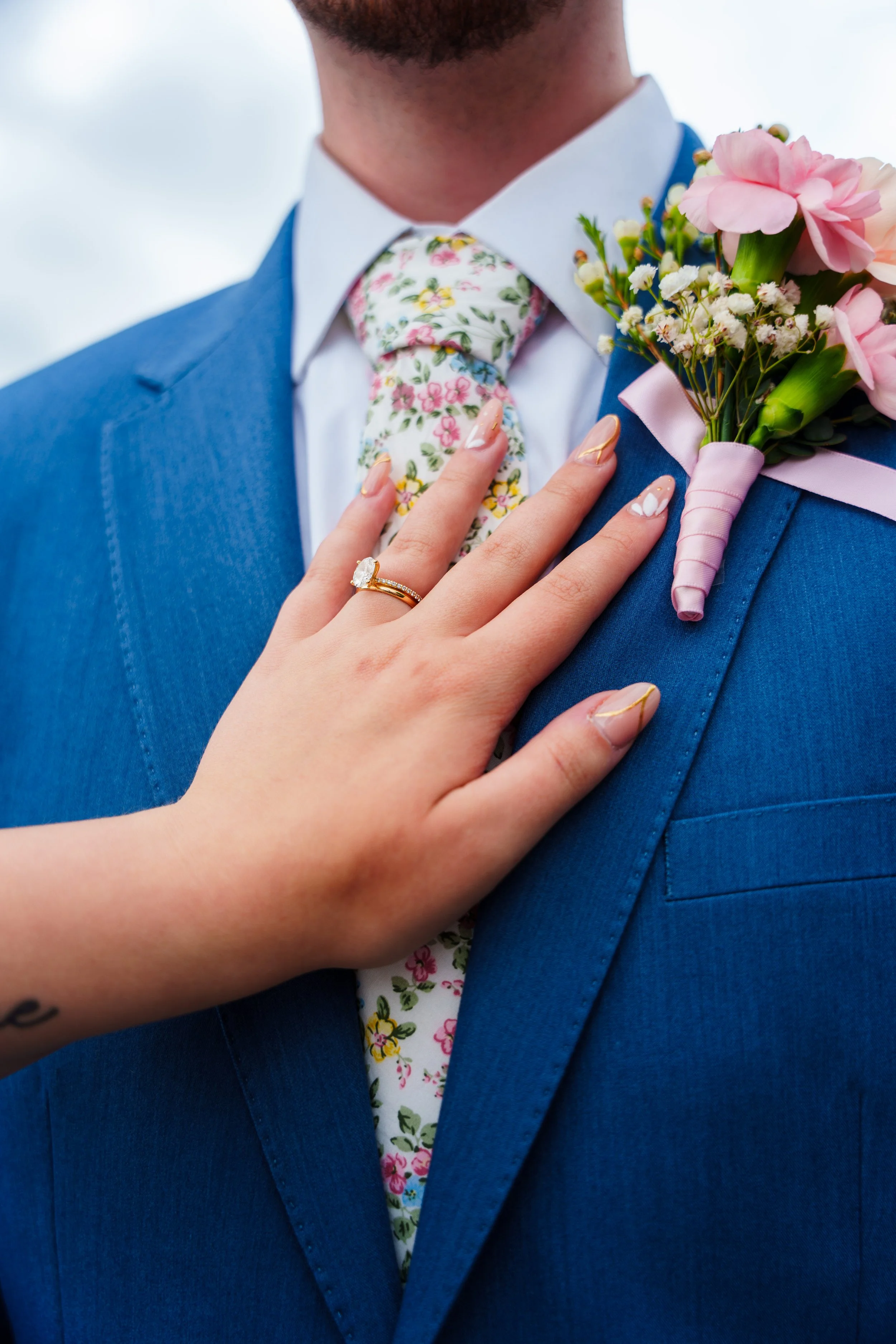 Close-up of a man in a blue suit with a floral tie and a white shirt, showing a pink wedding boutonniere on his lapel. A woman's hand with a wedding ring rests on his chest.