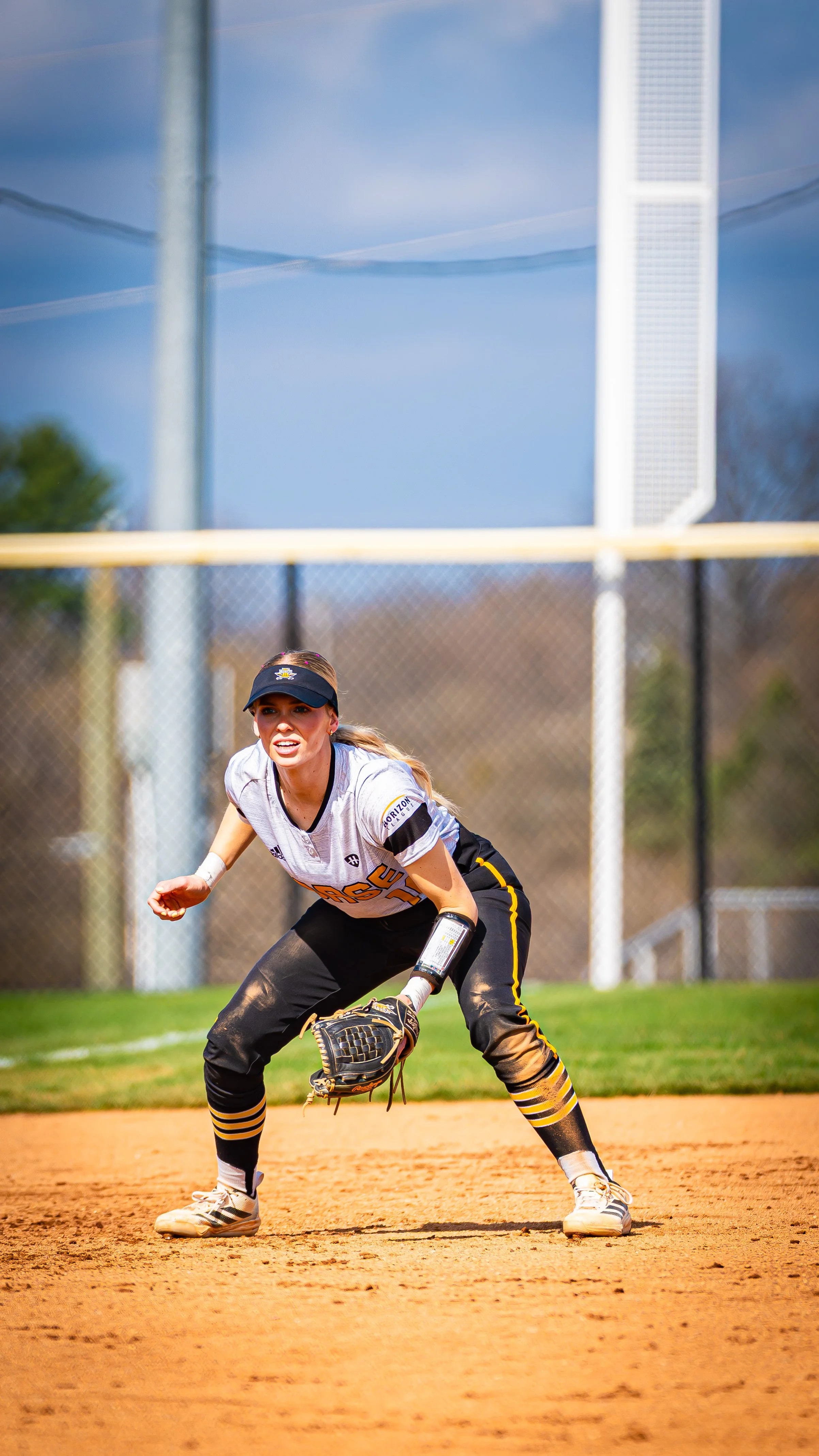 A female softball player in a white and black uniform with yellow accents is in a ready position, preparing to catch a ball on a softball field.