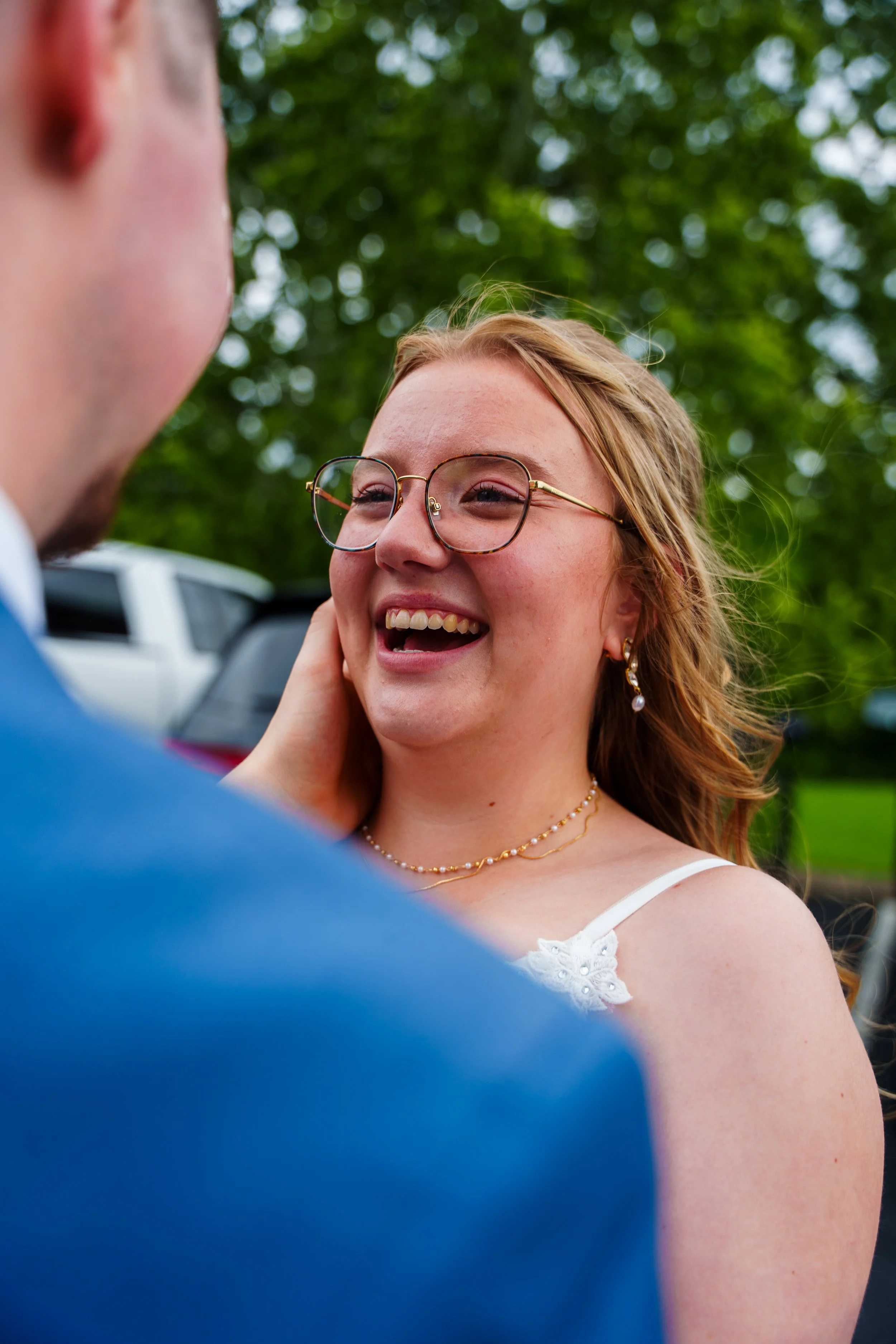 A woman with glasses, earrings, and a pearl necklace is smiling and looking into the eyes of a man who is touching her face. They are outdoors with green trees in the background.