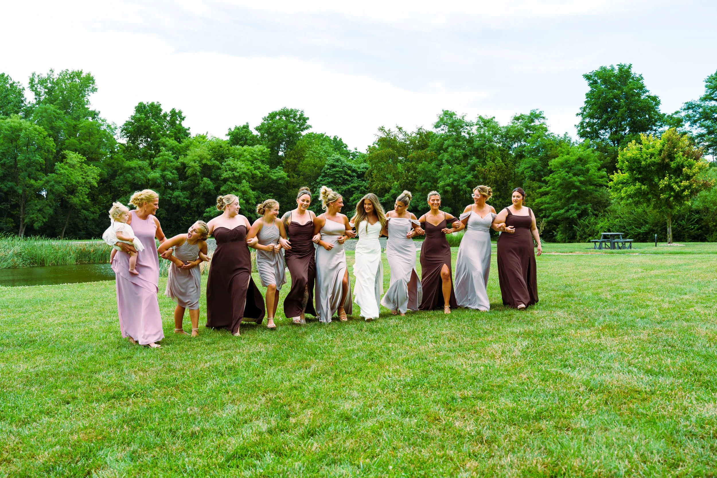 A group of women dressed in long gowns walking arm-in-arm across a grassy field with trees and a picnic table in the background.