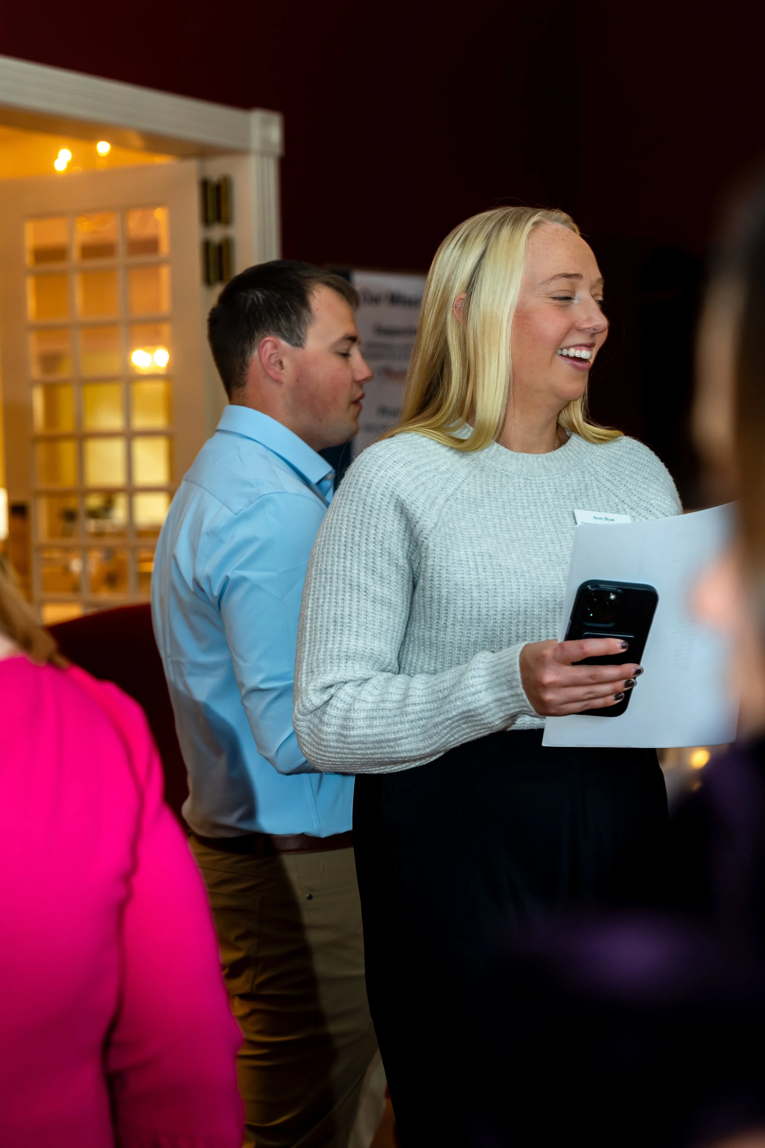 A woman with long blonde hair smiling as she looks at her phone and holds a paper, standing among a group of people.