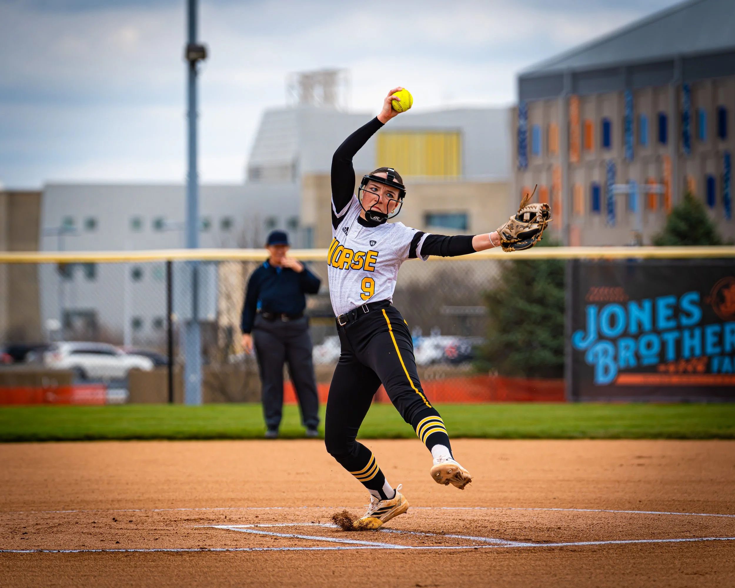 A female softball player in a gray and black uniform preparing to throw a yellow softball near a baseball field.