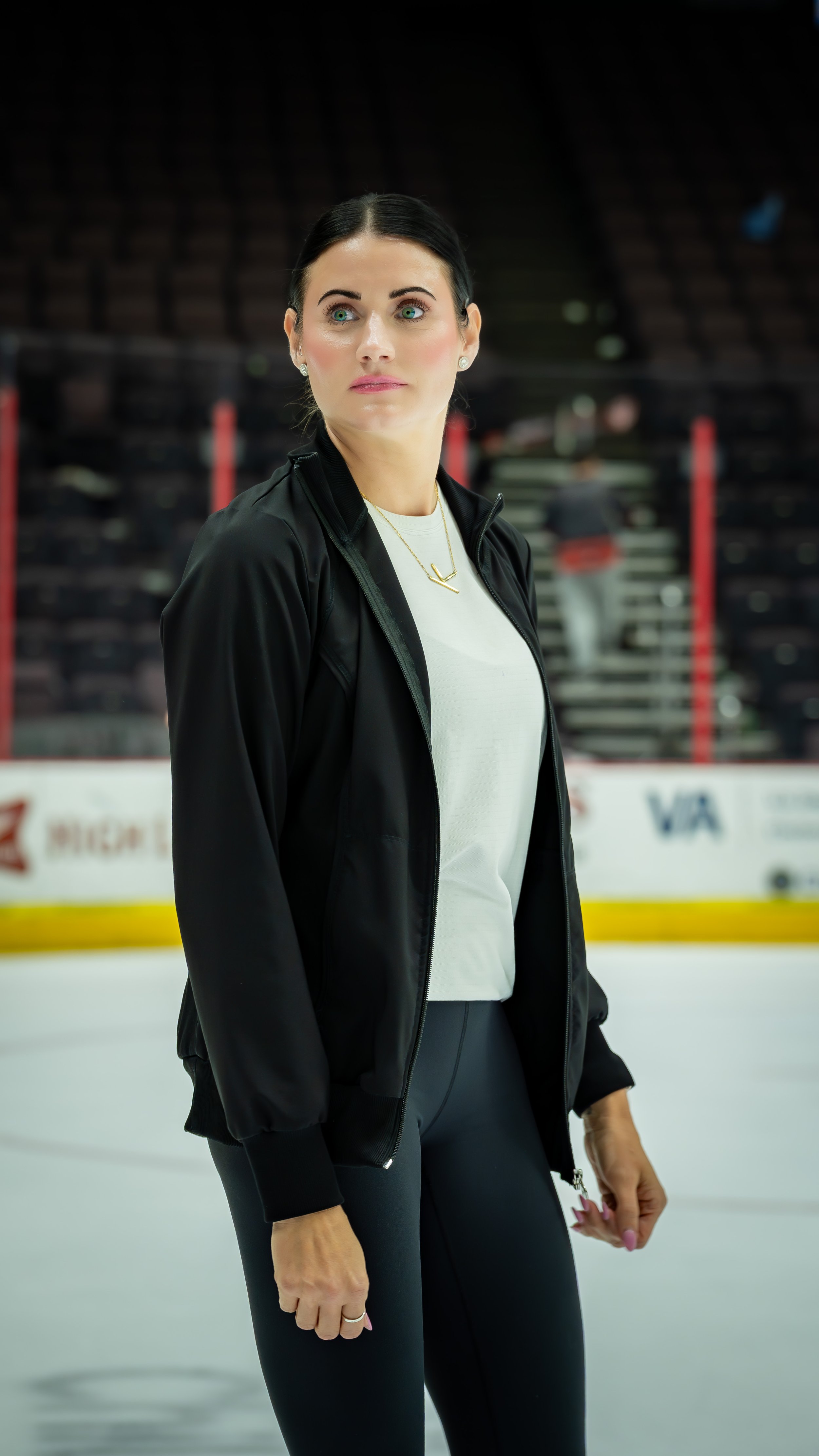 A woman standing on an ice rink, wearing a black jacket over a white shirt, with dark pants, in an indoor ice hockey arena.