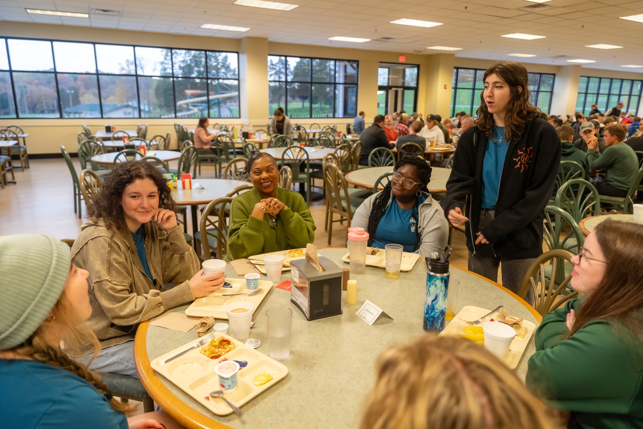 People sitting at tables inside a cafeteria or dining hall, eating food and engaging in conversation, with large windows showing trees and a water slide outside.