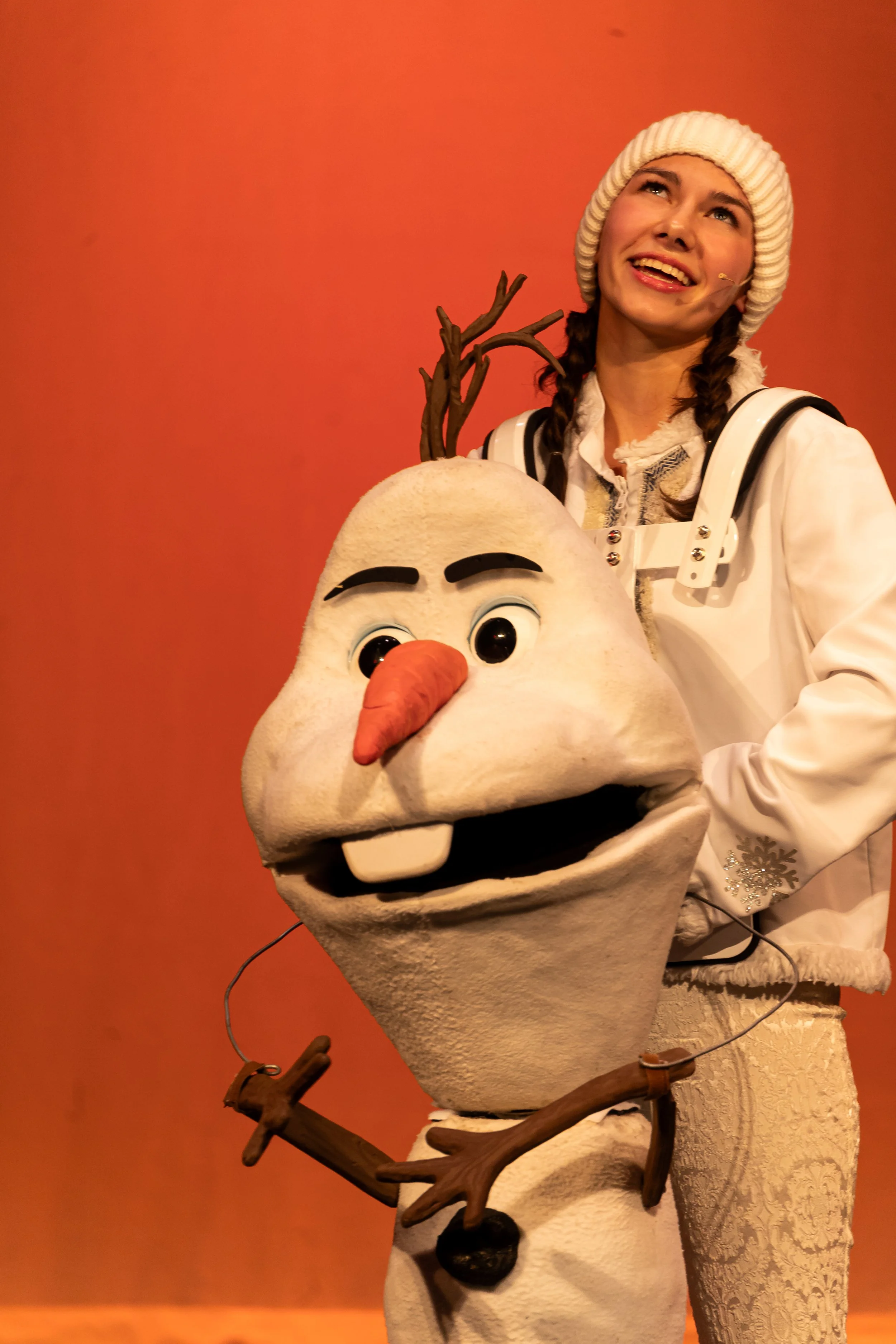 A person dressed as Olaf, the snowman character from Frozen, holding a branch and smiling, with a woman in a white knit hat and patterned white coat standing next to them against a red background.