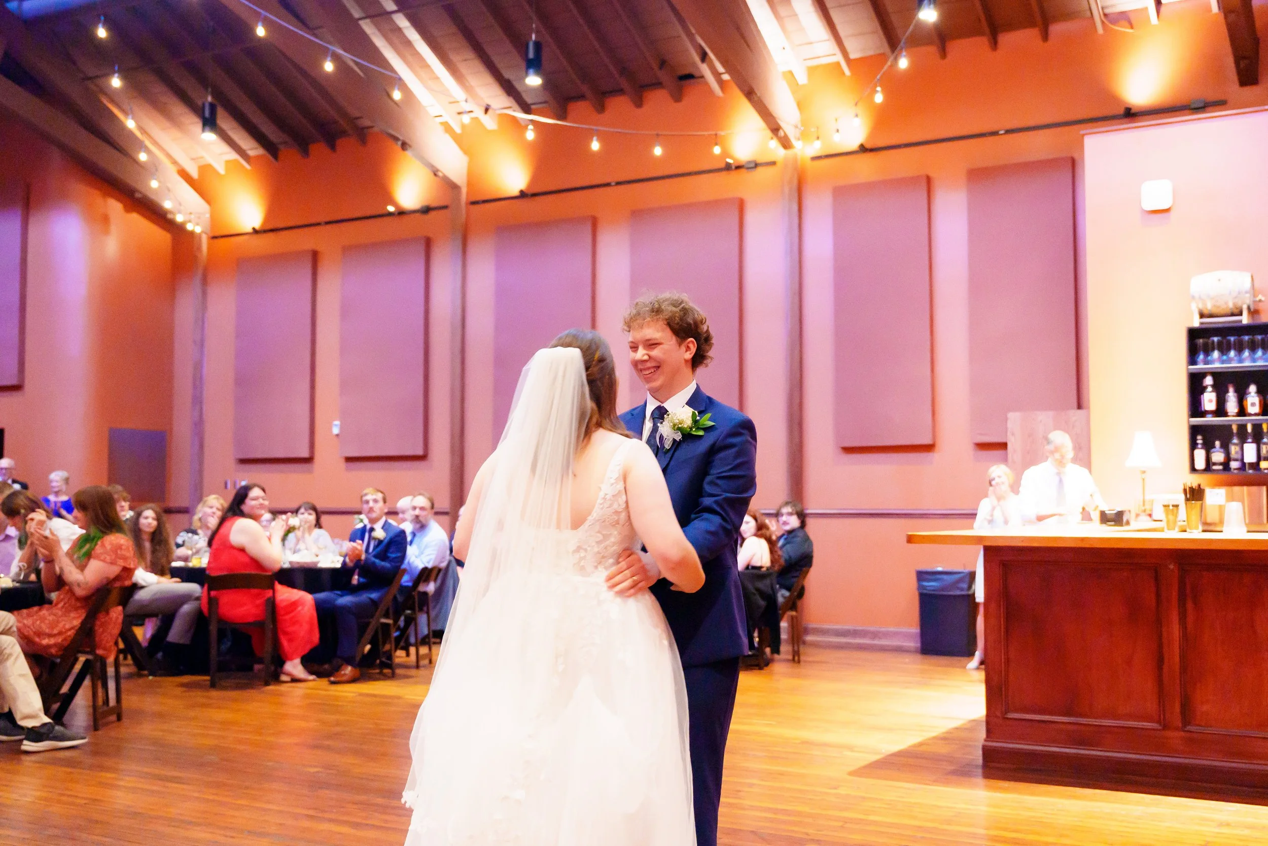 A bride and groom sharing a dance at a wedding reception, with guests seated at tables watching in a warmly lit room with string lights.