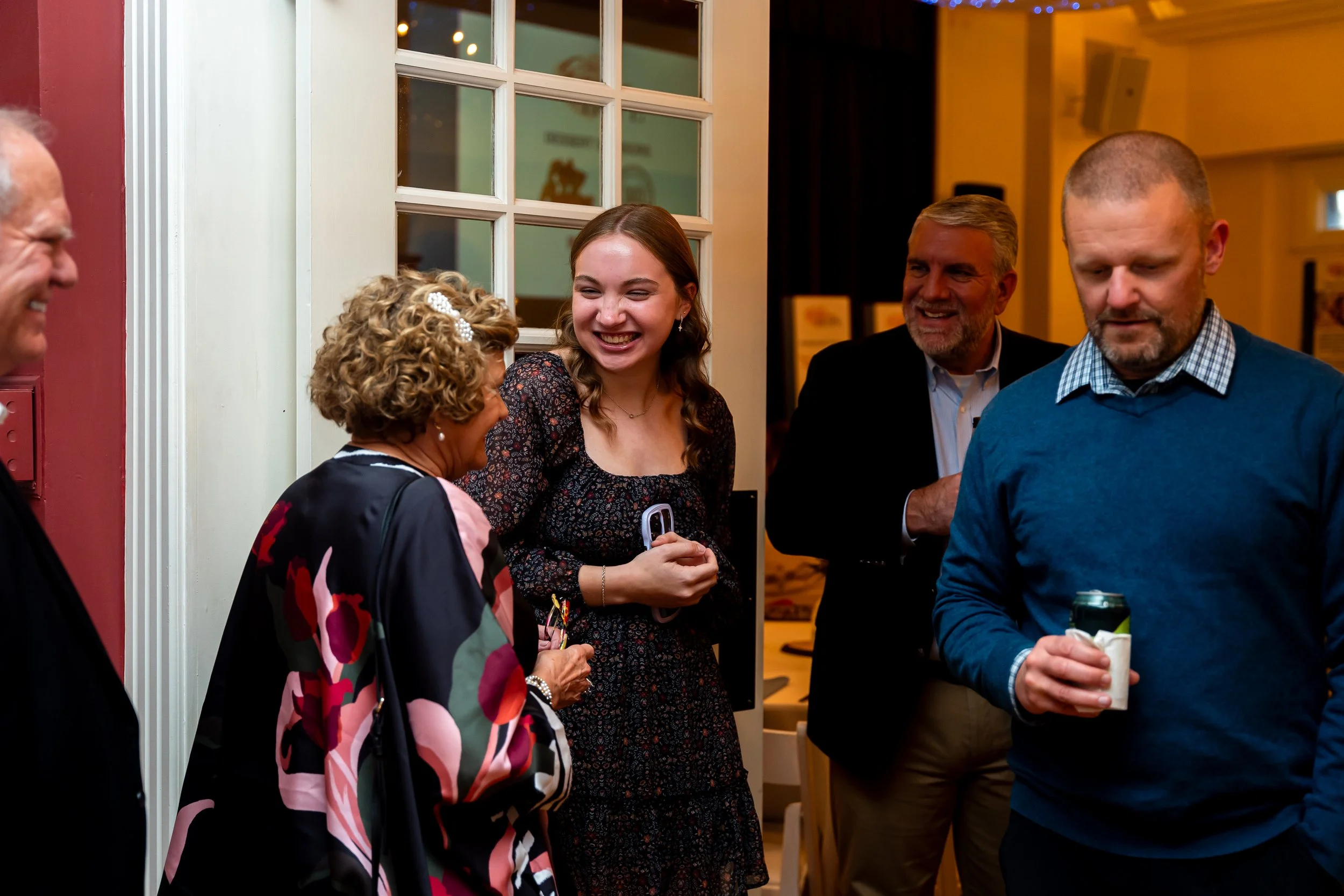 Group of five people talking at an indoor social gathering, smiling and holding drinks, near a windowed wall.
