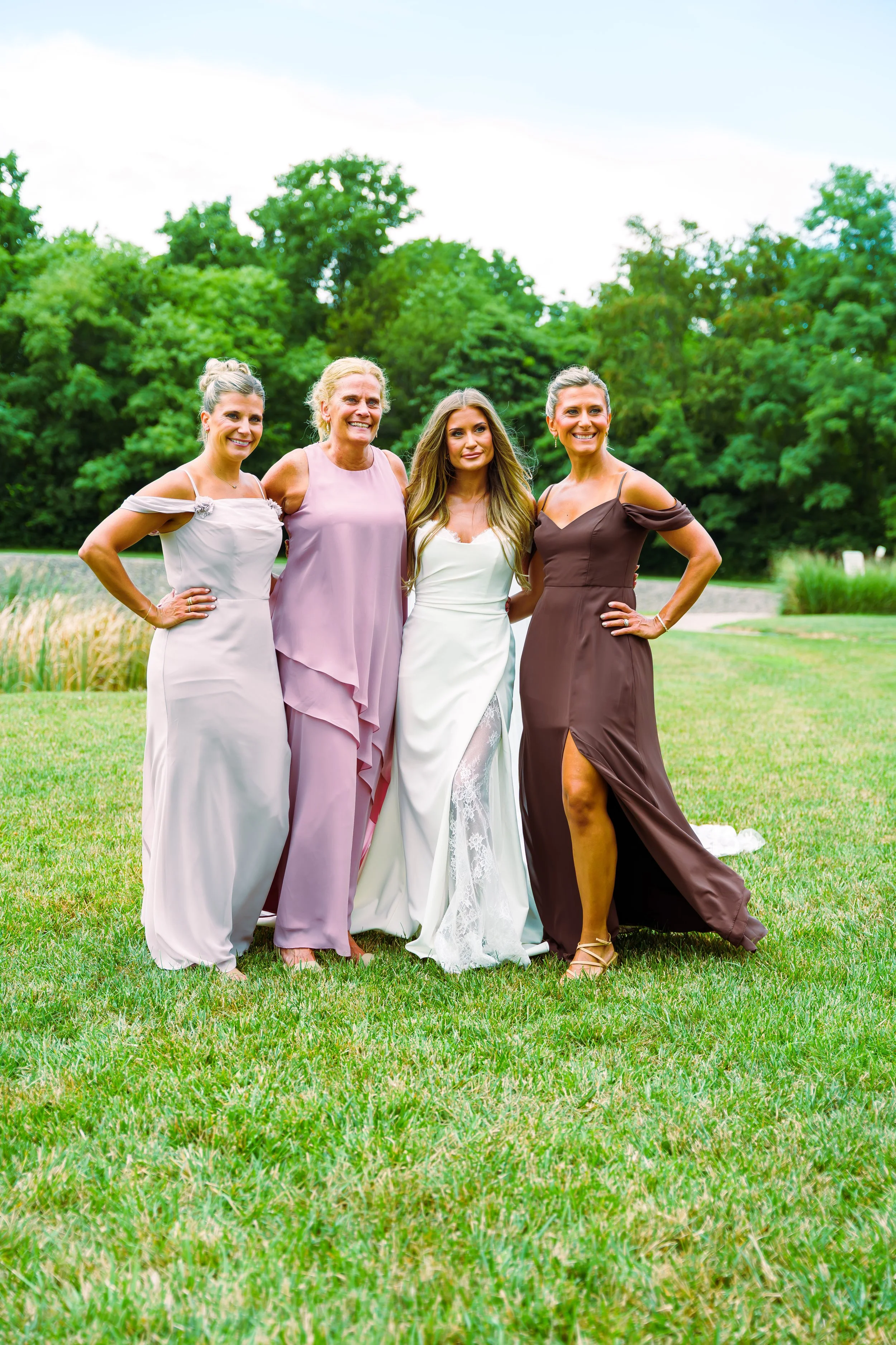Four women dressed in formal attire standing outdoors on a grassy area with trees in the background, smiling and posing for a photo.
