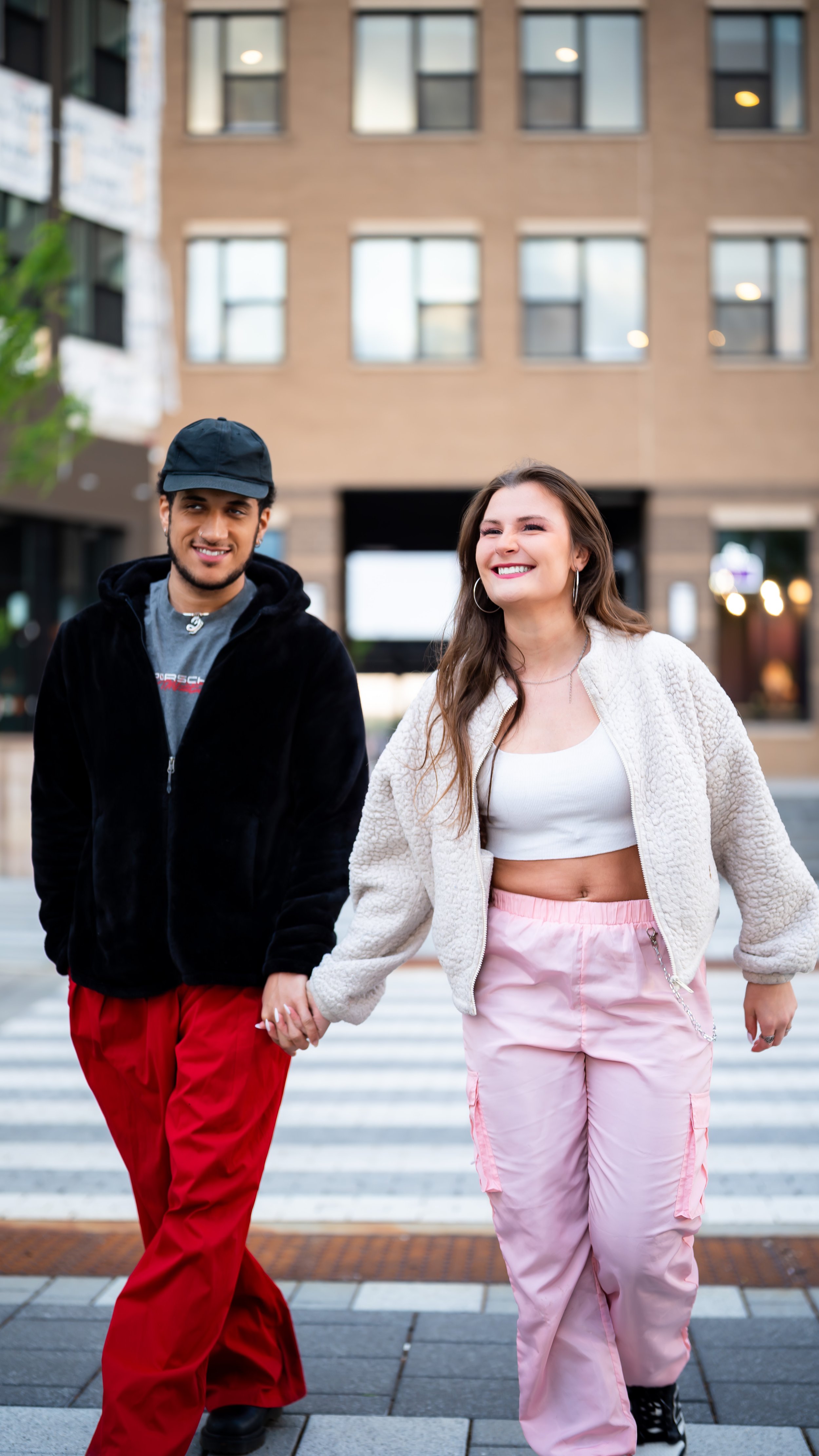 Two young women and a young man walking hand in hand across a city street, smiling, holding hands, with a building in the background.