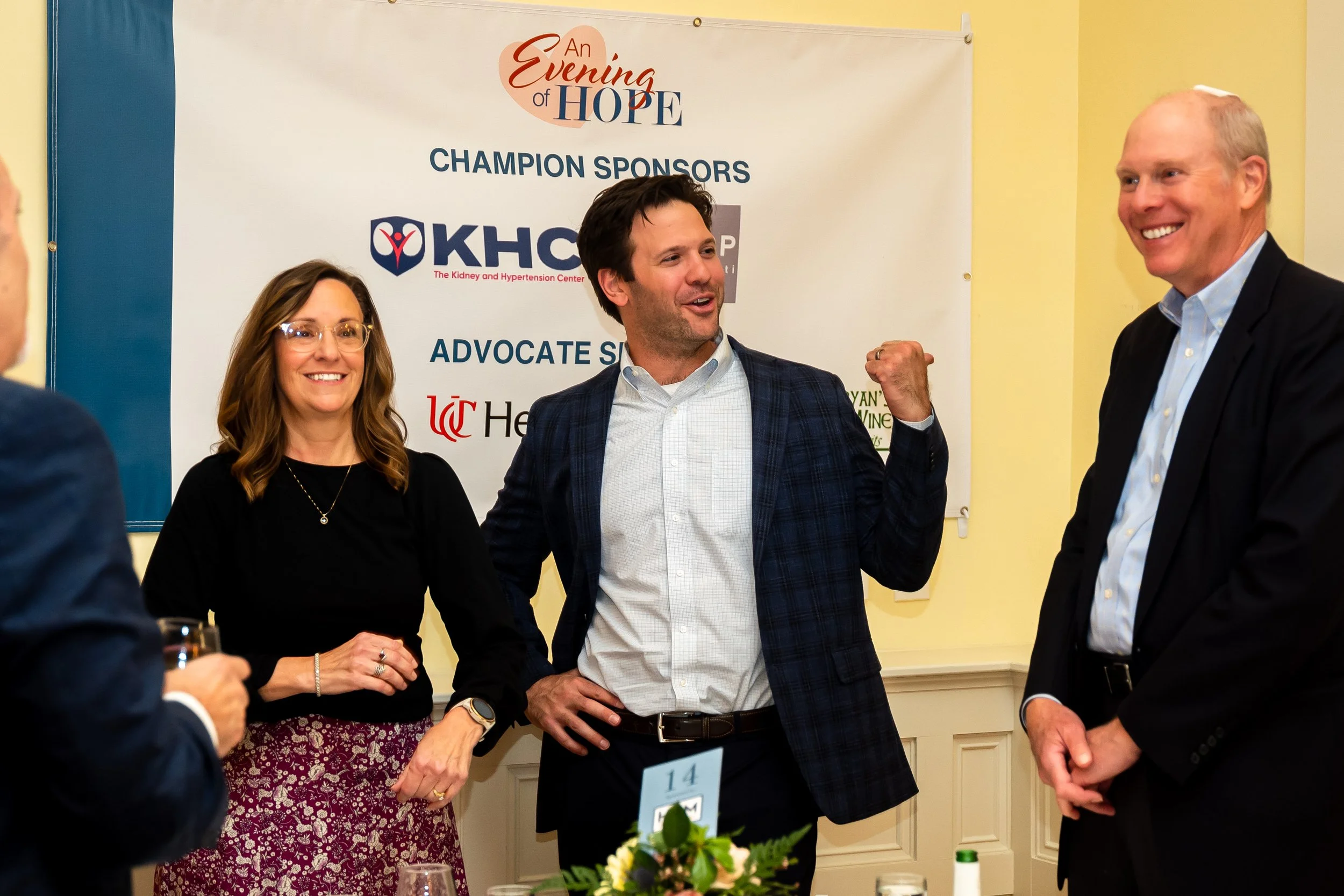 Three people are standing and talking at an event about kidney health, with a banner behind them that reads "An Evening of Hope" and sponsored by KHC. The woman on the left has long brown hair and glasses, the man in the center has dark hair and is g
