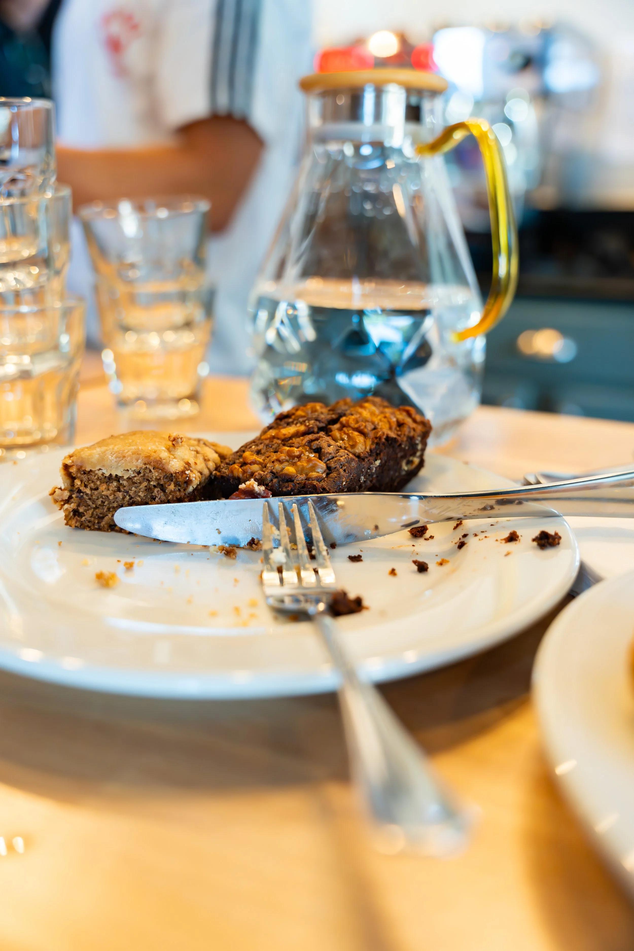 Partially eaten plate containing two pieces of dessert, one resembling a cake and the other a nut-topped bar, on a table with a fork and knife, in front of a glass pitcher of water, with glasses and blurred background.