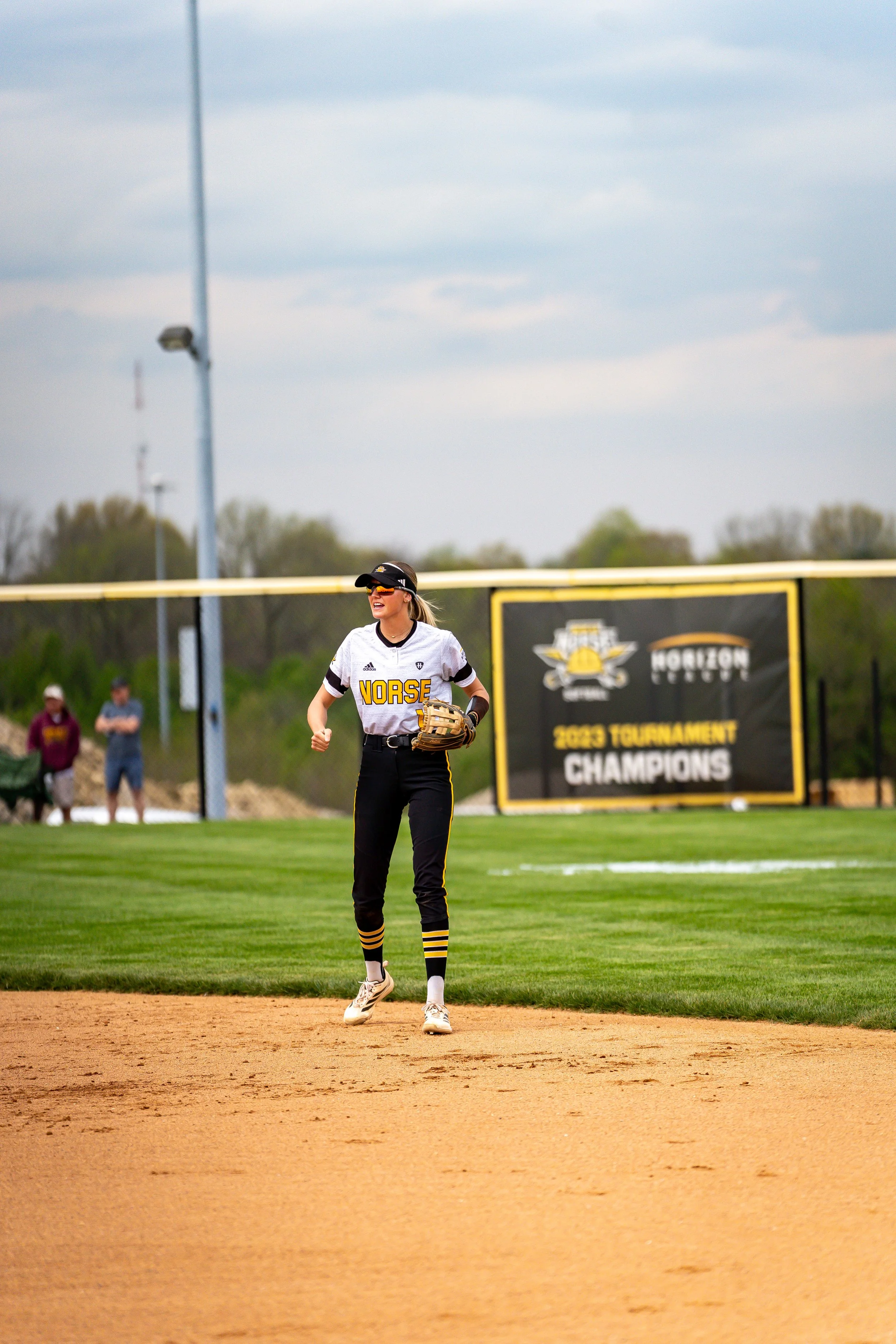 A female softball player standing on the field with a black hat, sunglasses, white jersey with black and yellow accents, black pants with yellow stripes, and cleats. She is smiling and holding a glove on her left hand. In the background, there is a y