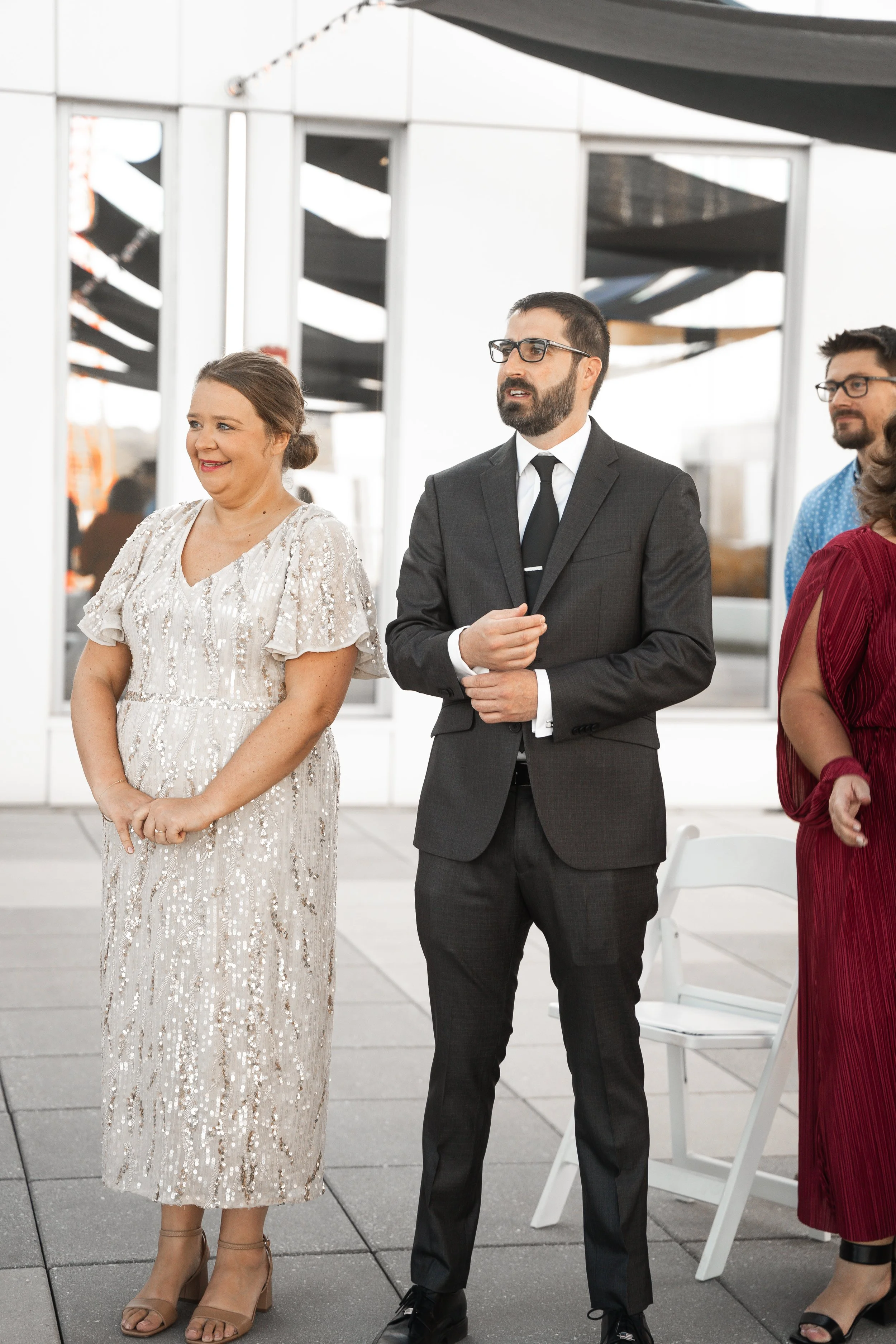 People dressed in formal attire, standing outdoors near white chairs and large mirrors with a modern architectural background.