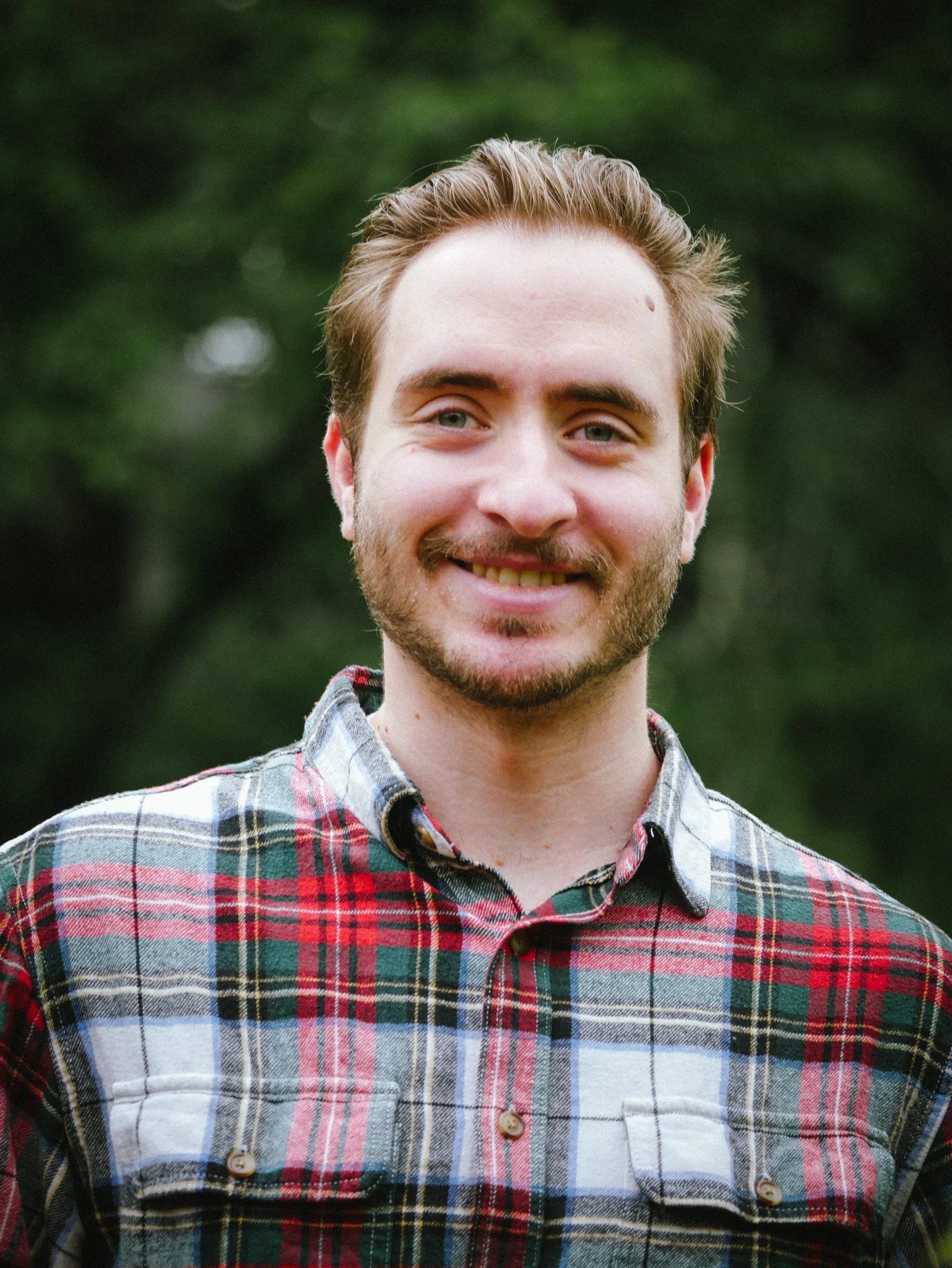 Close-up of a smiling young man with light skin, light brown hair, and a beard wearing a plaid shirt, standing outdoors with a blurred green background.