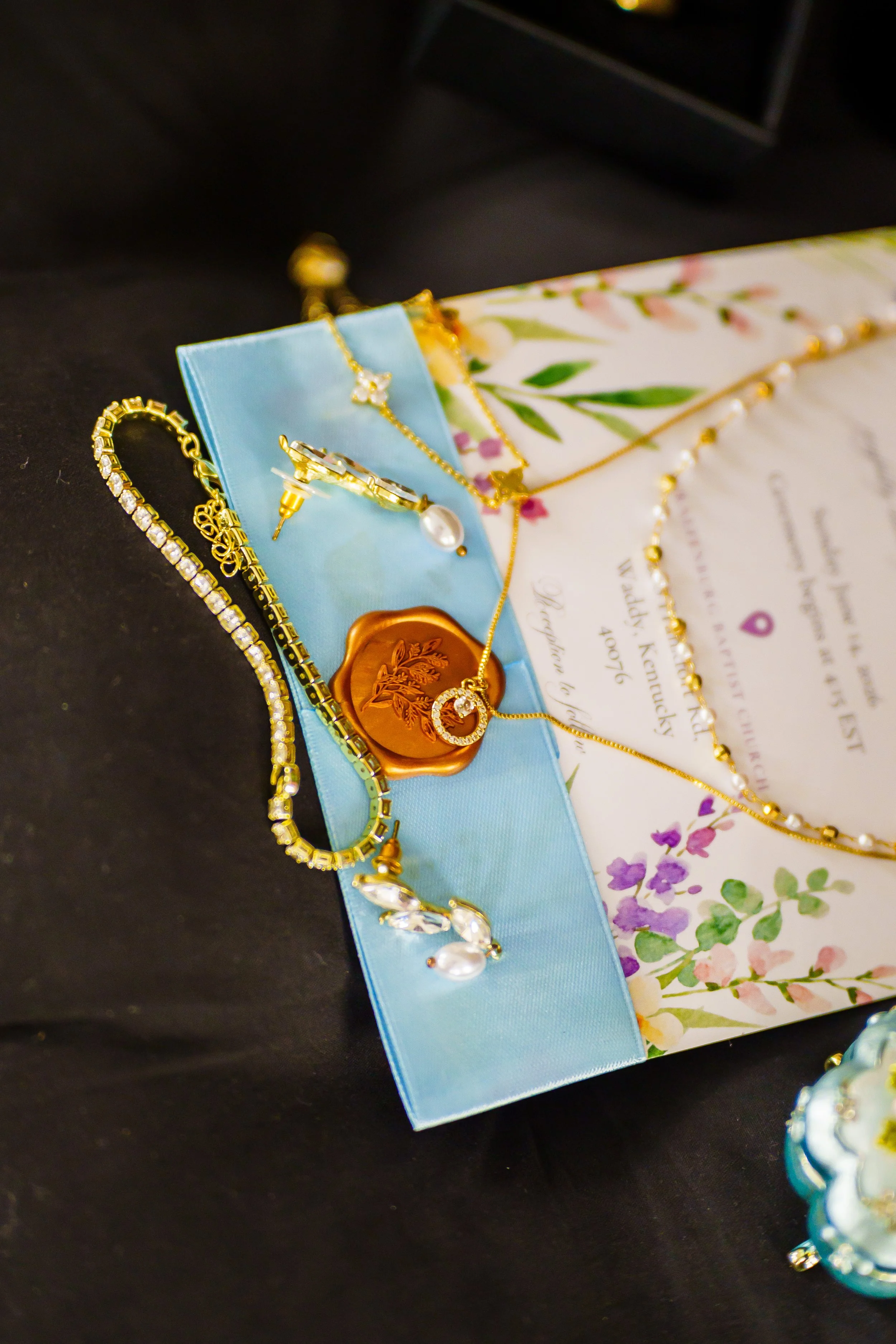 Close-up of jewelry displayed on a decorative floral paper with a light blue ribbon, including gold and diamond necklaces, earrings, and a pendant.
