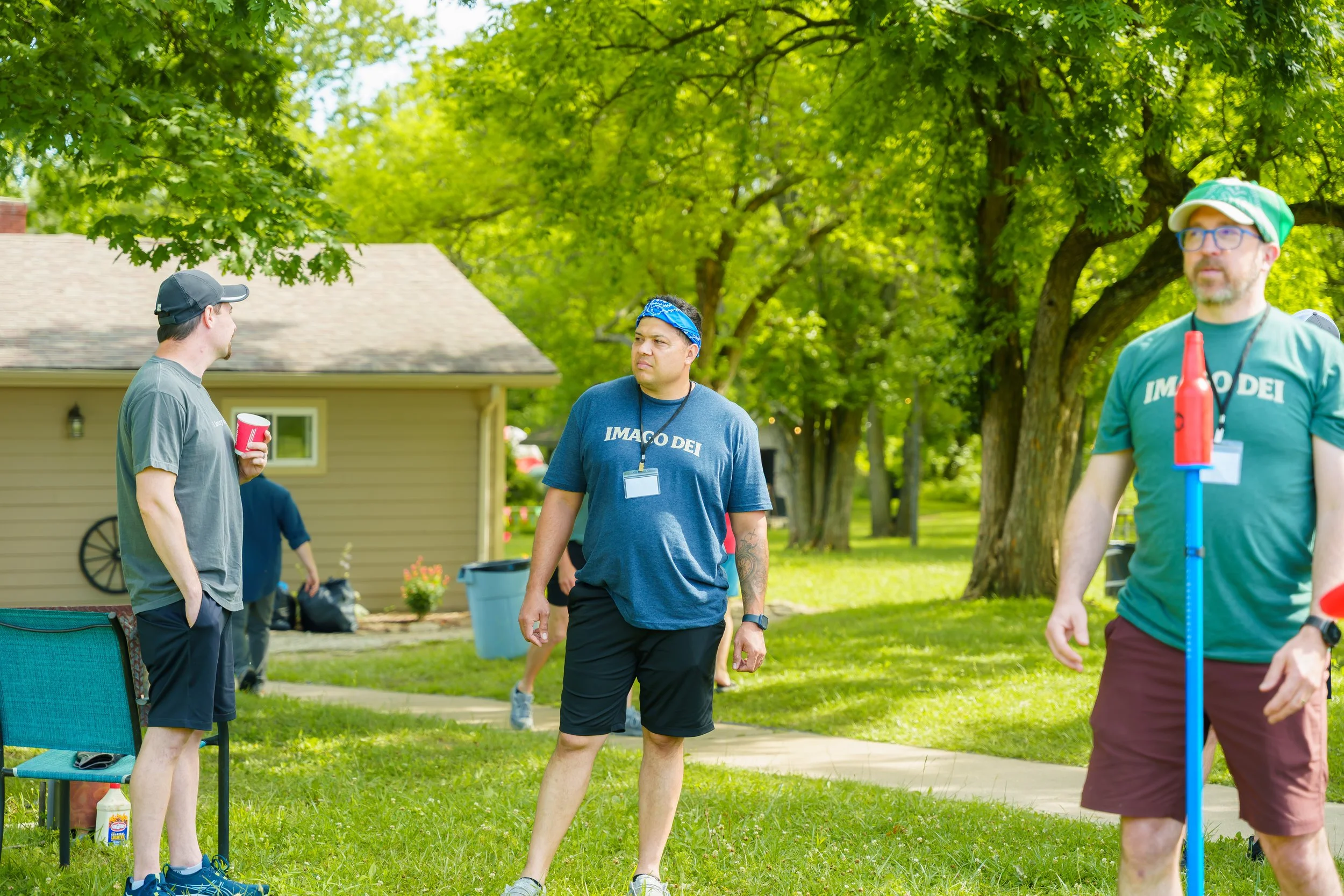 Three men are standing outdoors in a park with green trees and a house in the background. They are engaged in conversation. One man on the left holds a red cup, the man in the middle wears a blue bandana and a t-shirt that says 'IMAGO DEI', and the m