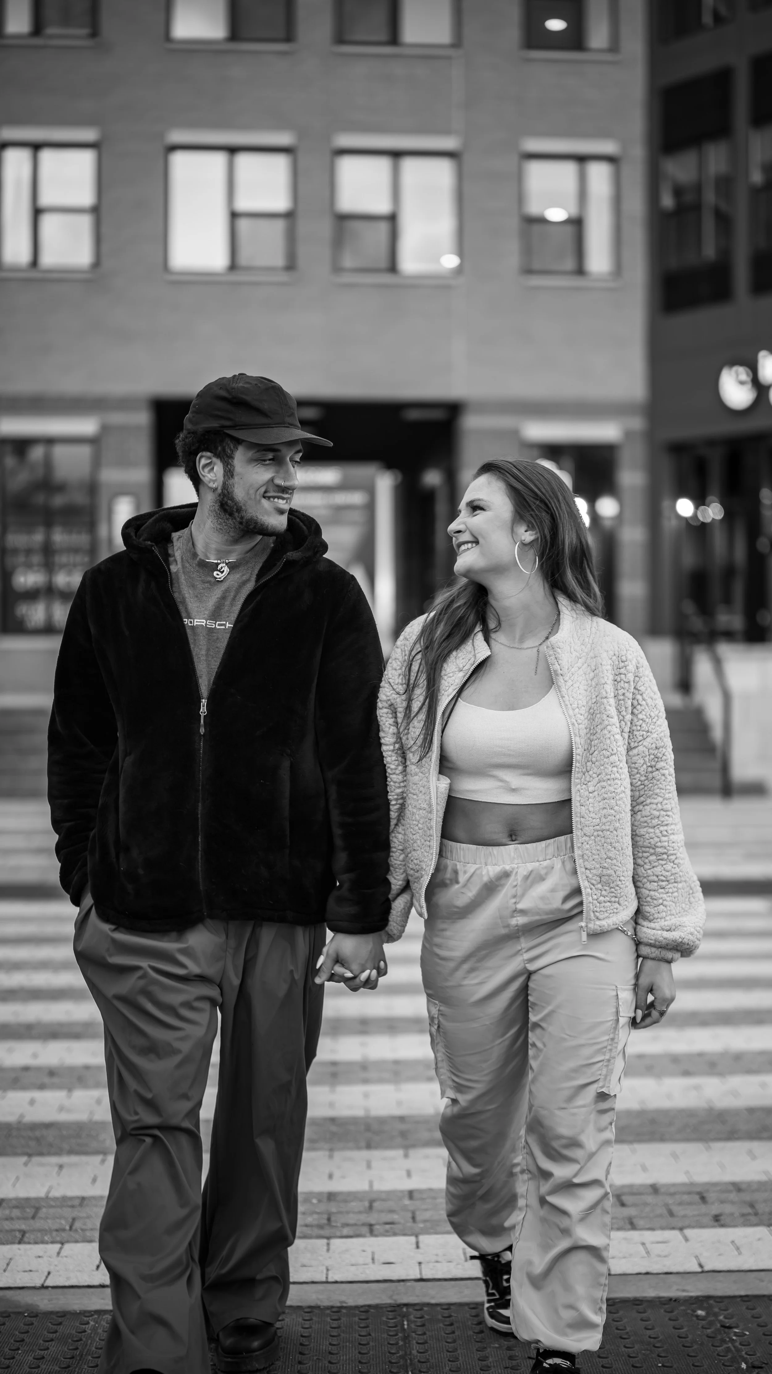 A young man and woman walk hand in hand across a crosswalk in an urban environment, smiling at each other. The man is wearing a cap, hoodie, and cargo pants, while the woman is dressed in a crop top, jacket, and cargo pants with sneakers.