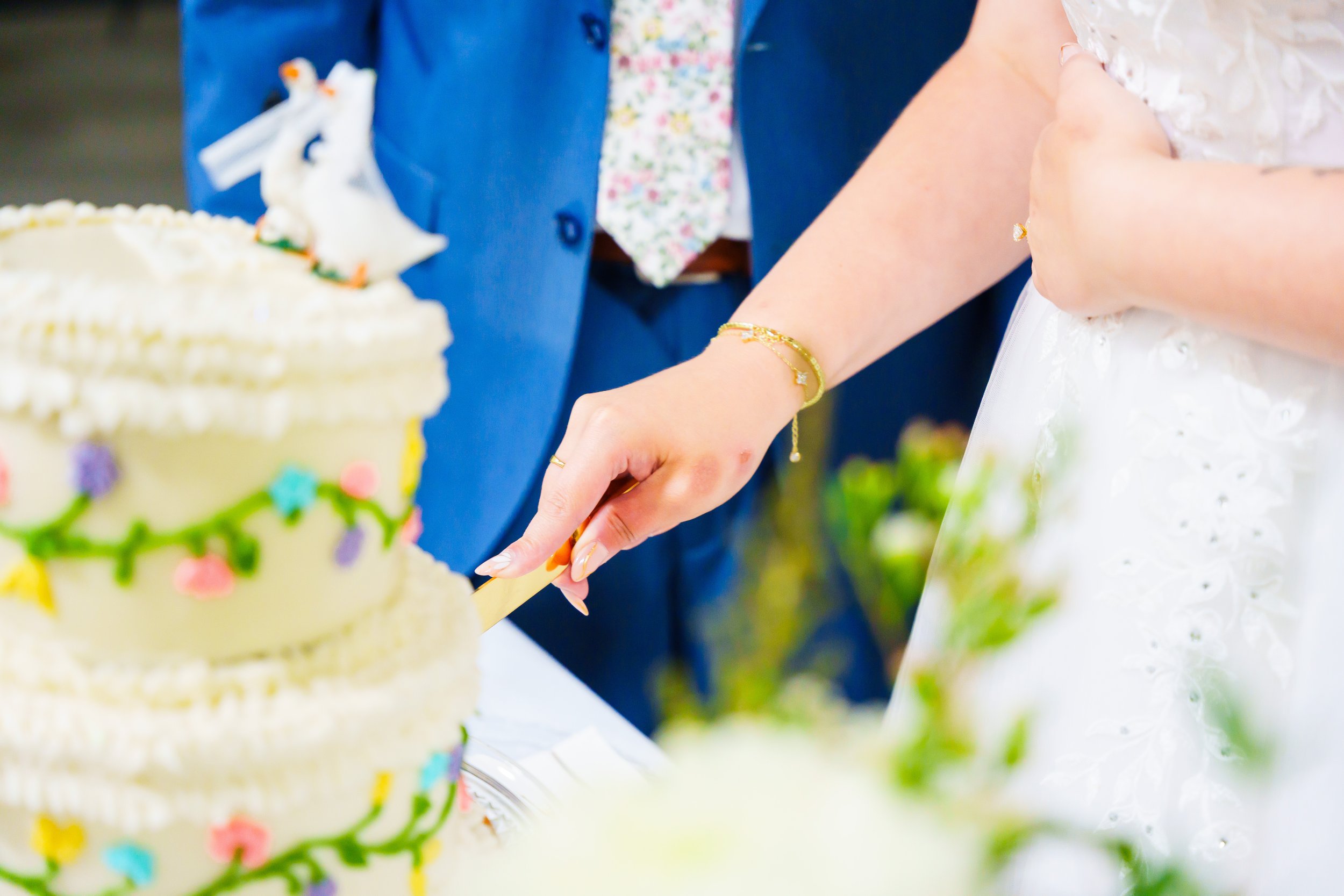 Close-up of a person cutting a decorated birthday cake with colorful icing and flowers, wearing a white dress and a gold bracelet.
