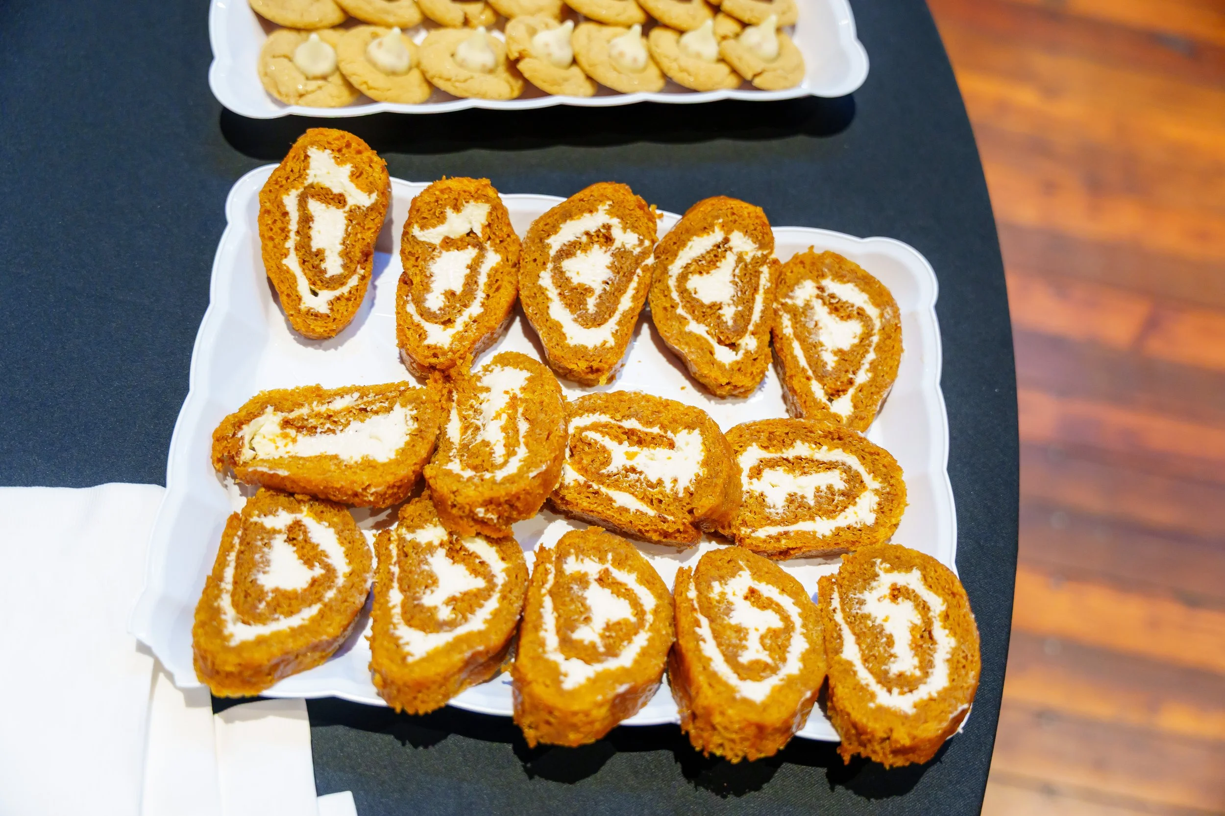 Cake roll slices with cream filling on a white plate, and cookies with cream topping on a white tray, placed on a dark table.