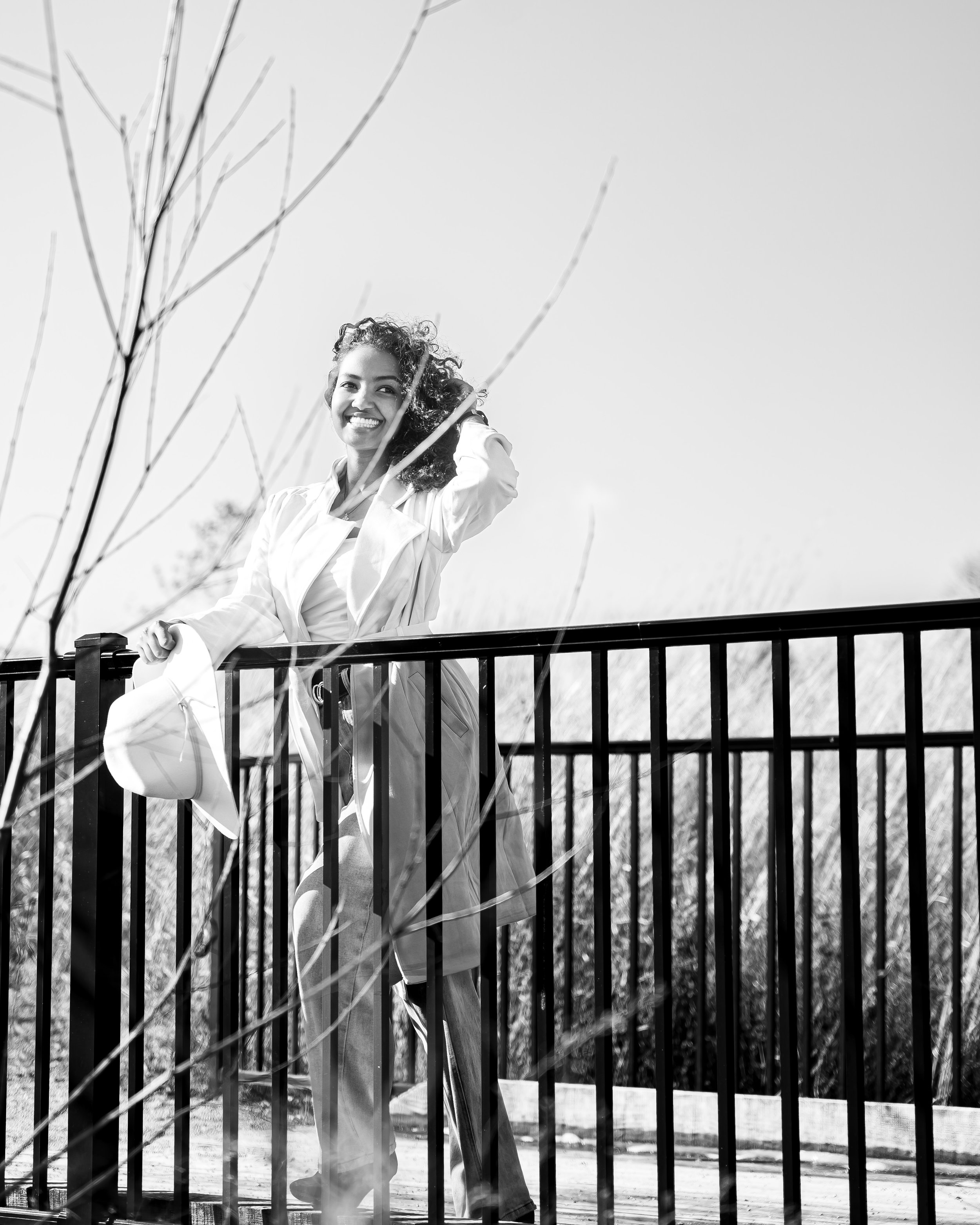 A young woman with curly hair smiling, wearing a light-colored blazer and pants, standing on a balcony with a railing outdoors. She holds a hat in her left hand, and her right hand is touching her hair. Bare tree branches are visible in the foregroun