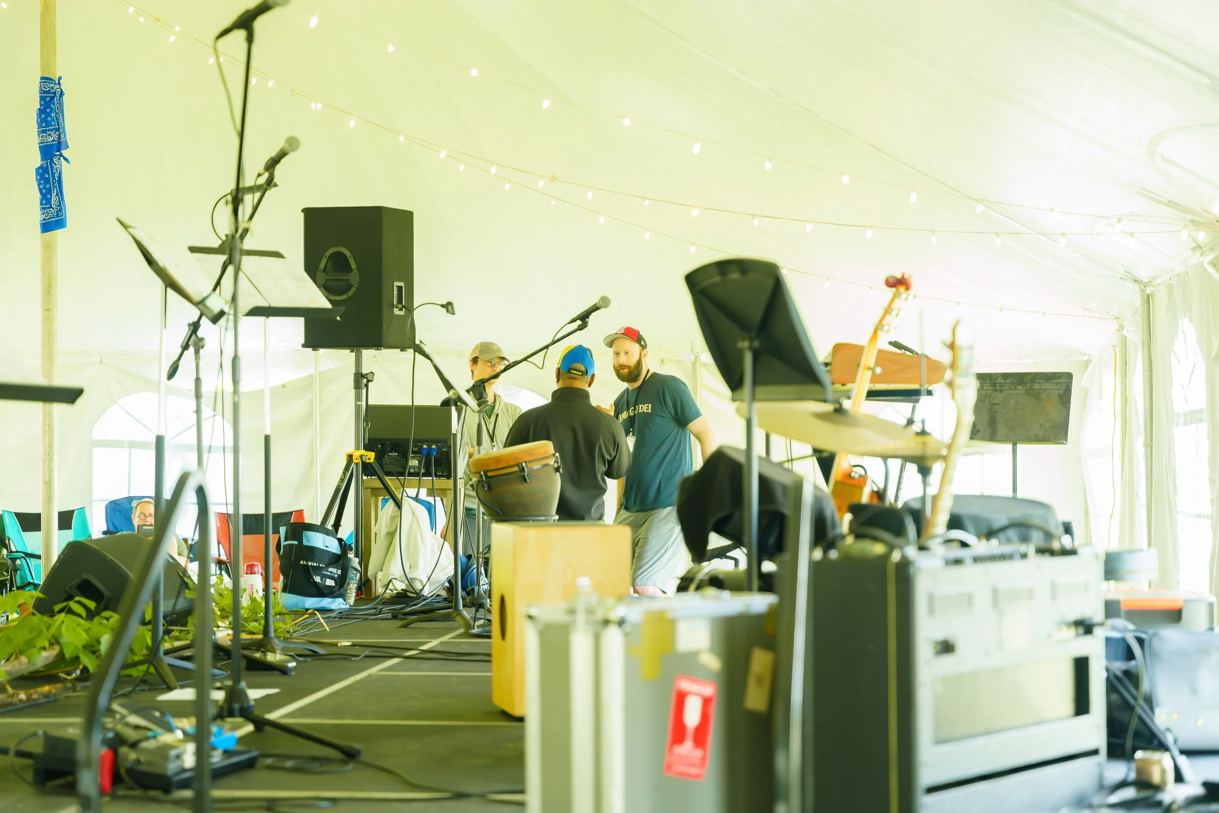 People setting up musical equipment and instruments inside a large tent with string lights and various stage gear.