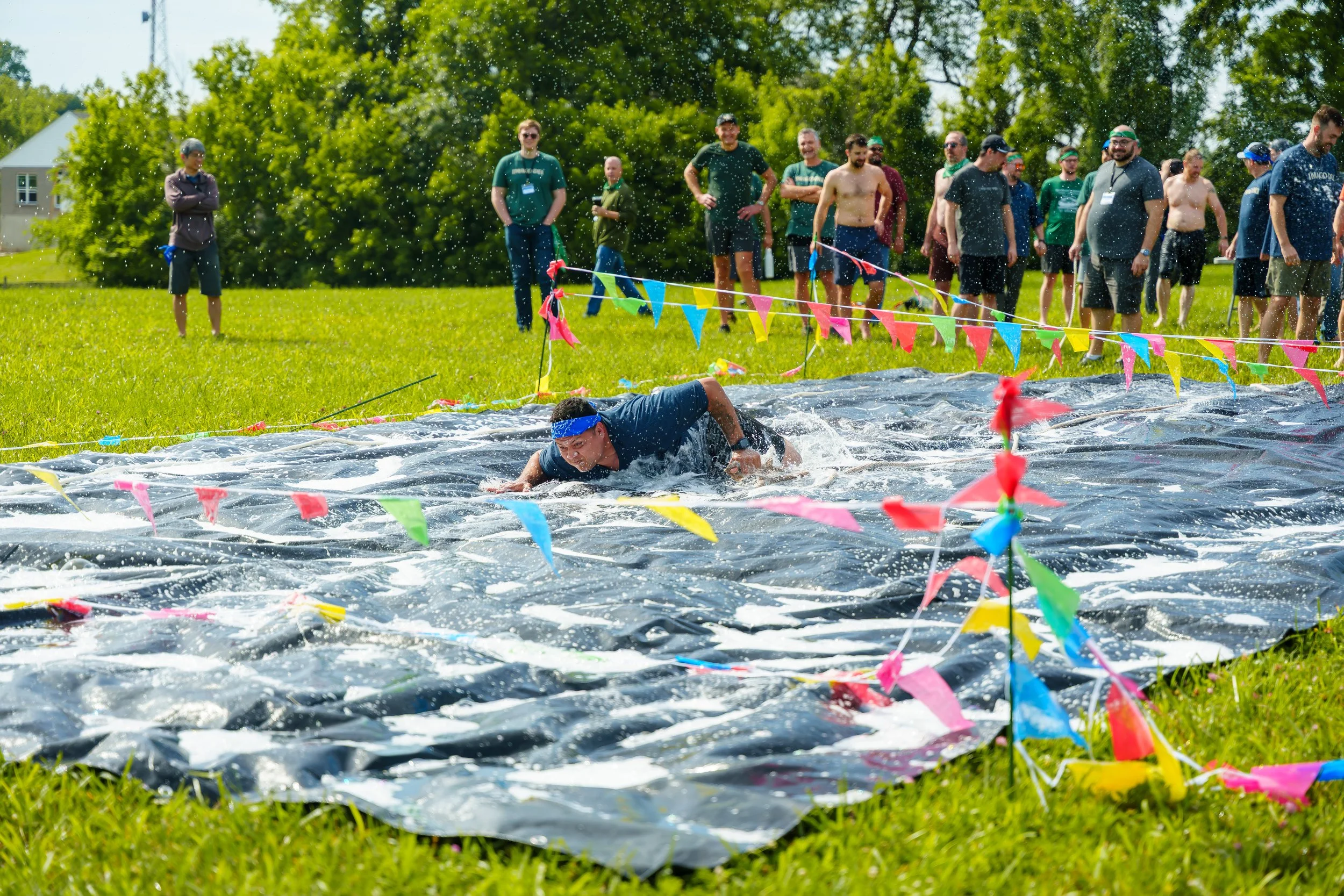 A person crawling on a black tarp in a grassy outdoor area, surrounded by colorful triangular flags, with a group of people standing and watching nearby.
