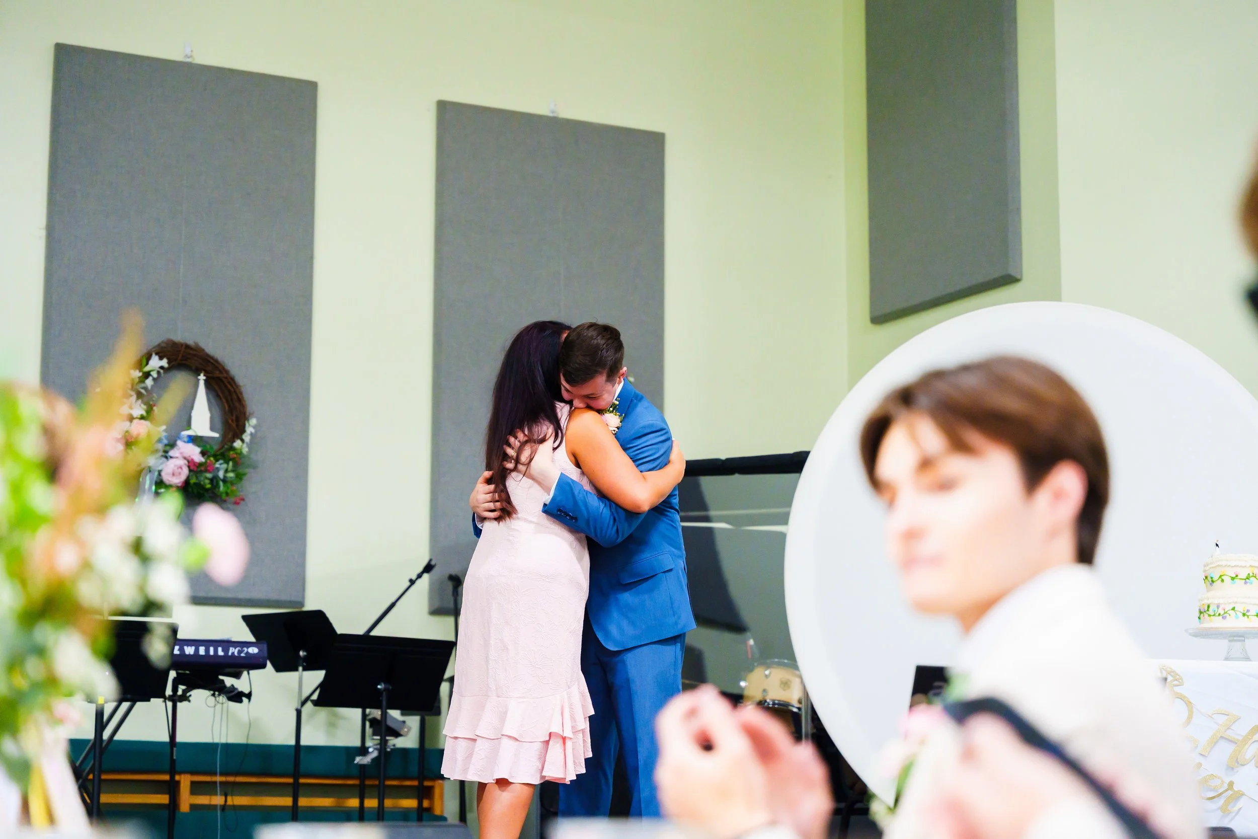 A couple hugging at a wedding or celebration, with the woman wearing a white dress and the man in a blue suit. In the foreground, a person with brown hair is partially visible, and a decorated cake is on a table. The background shows a light green wa
