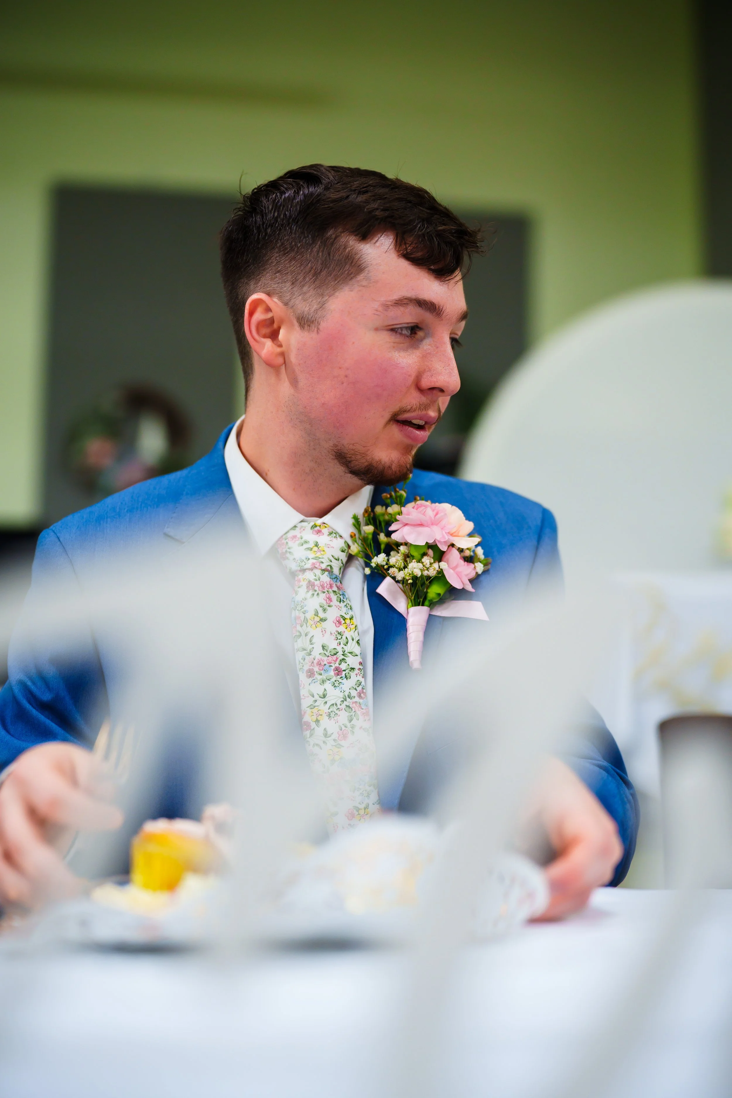 A young man in a blue suit with a floral tie and a boutonniere is participating in a celebration, possibly a wedding, holding a piece of paper or cards. He has short dark hair and a slight beard, and the background features a green wall and a mirror.