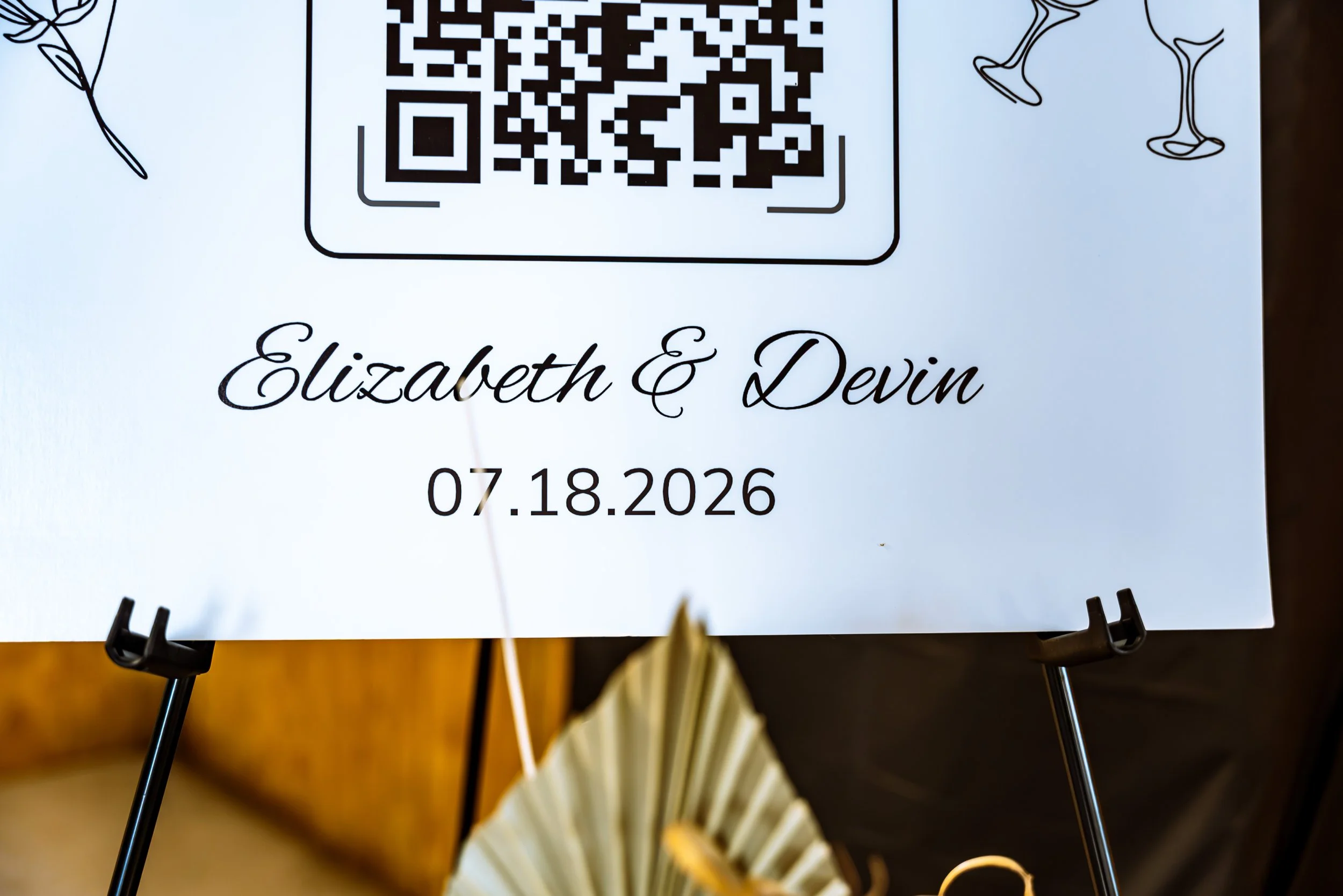 A white sign with black script font displaying the names Elizabeth & Devin and a date of 07.18.2026. There is a QR code at the top of the sign, and decorative floral illustrations in the corners. The sign is supported by a black stand.