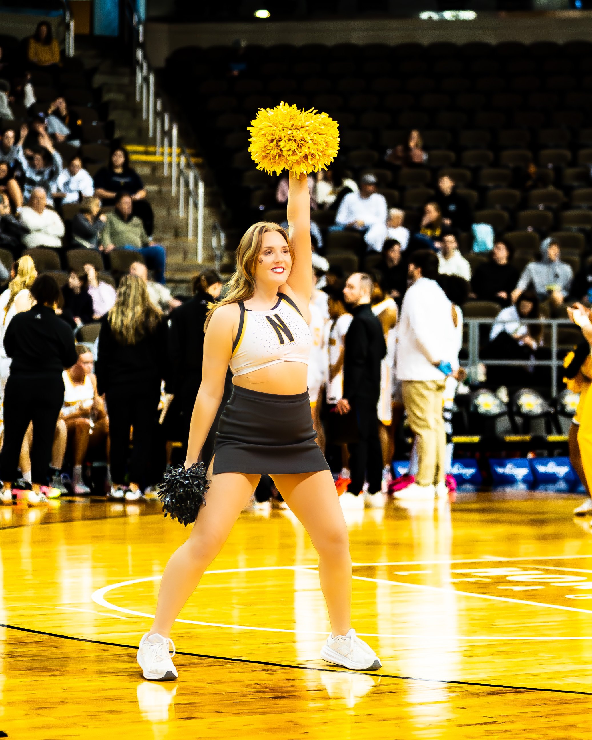 A cheerleader in a black and white uniform holding yellow pom-poms, standing on a basketball court during a game, with spectators seated in the background.