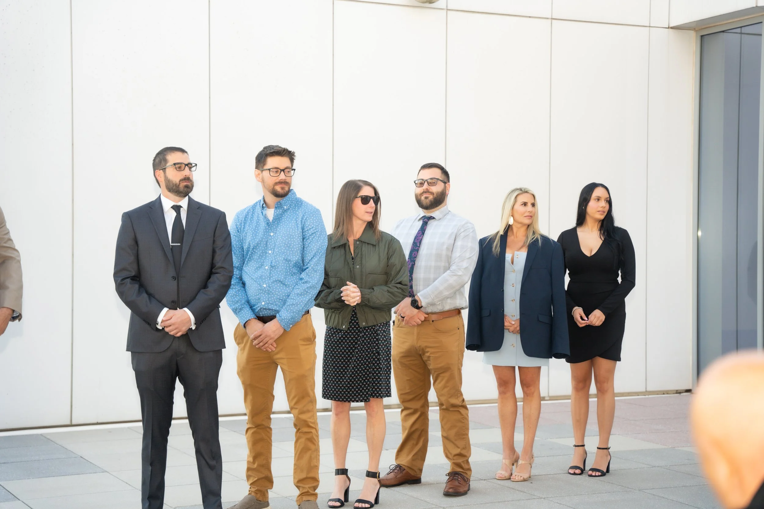 Group of six diverse people standing outside against a white wall, dressed in business casual and formal attire, some wearing sunglasses, appearing to wait or watch something.