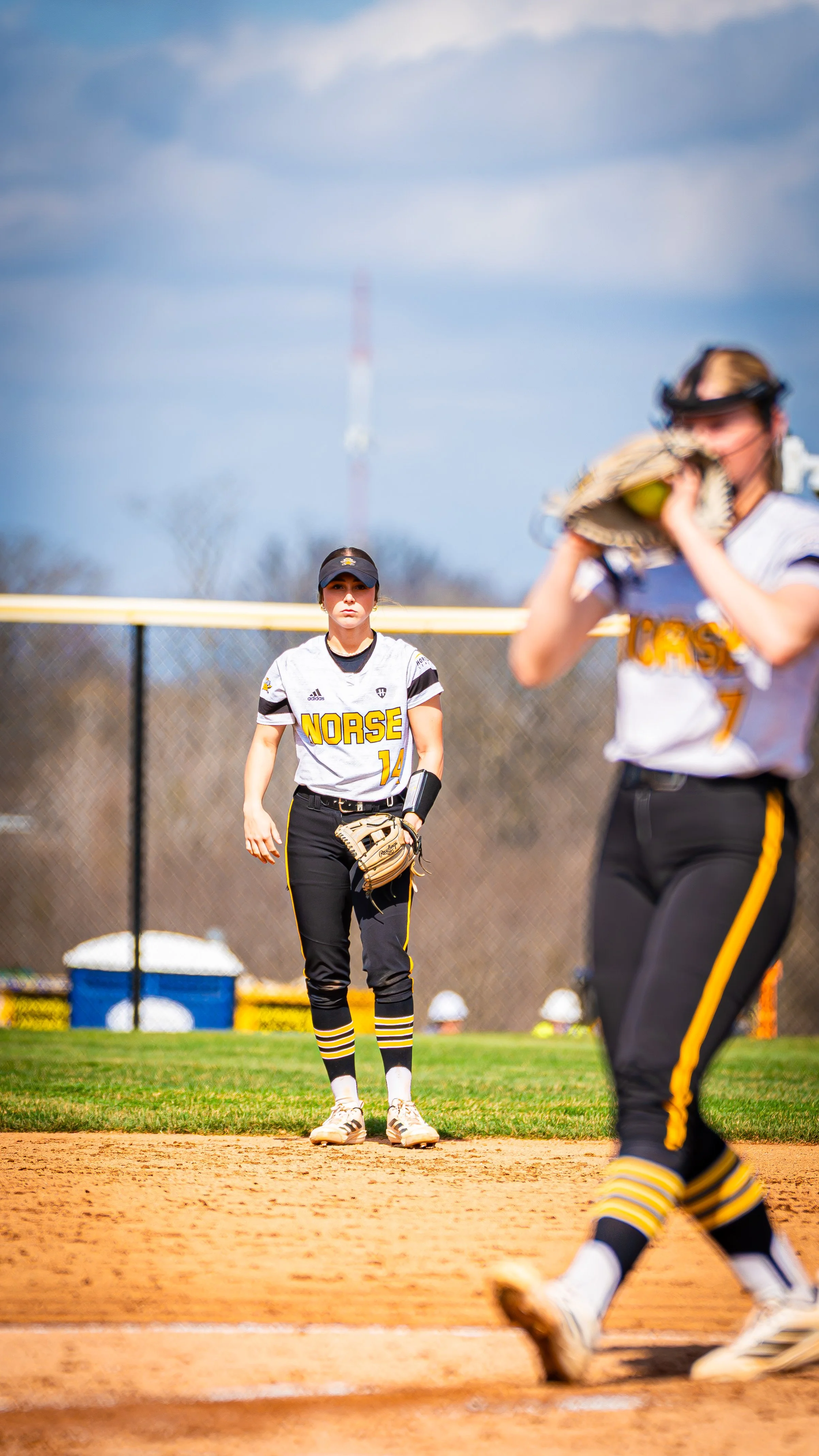 A softball player standing on the field with her glove on her left hand, wearing a white and black uniform with 'Norse' written on it, and a black cap, during a game. Another player is in the foreground, with part of her body and glove visible, and a