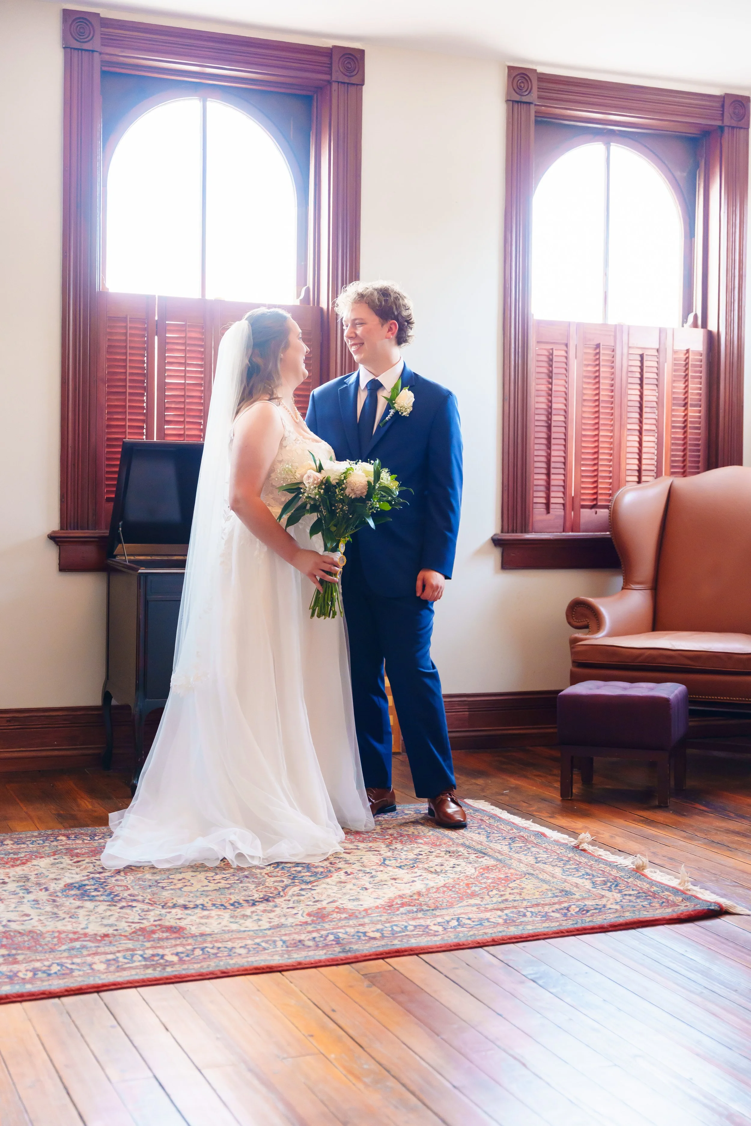 A bride and groom are standing indoors, looking at each other and smiling, during their wedding ceremony. The bride wears a white wedding dress and veil, holds a bouquet, and the groom wears a blue suit with a white shirt and blue tie. They are in fr