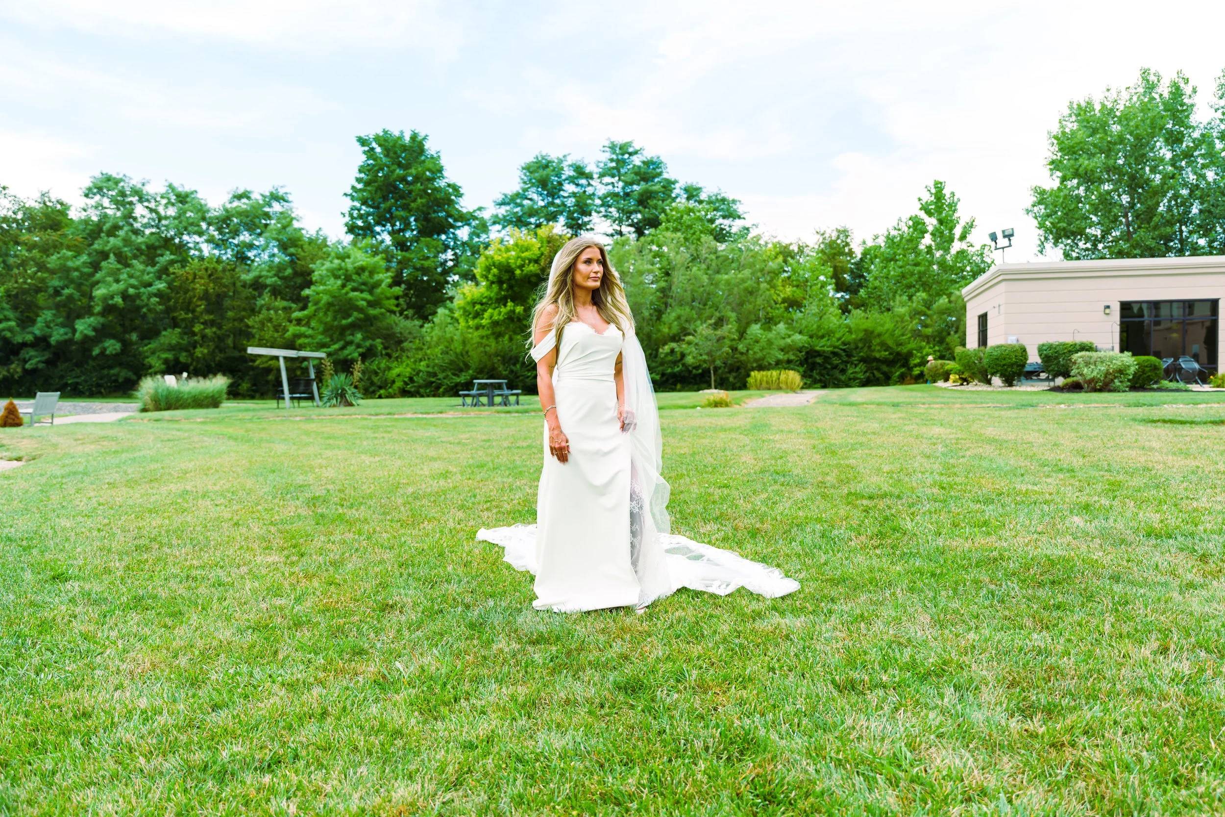 A woman in a white wedding dress standing on a green lawn with trees and a building in the background.