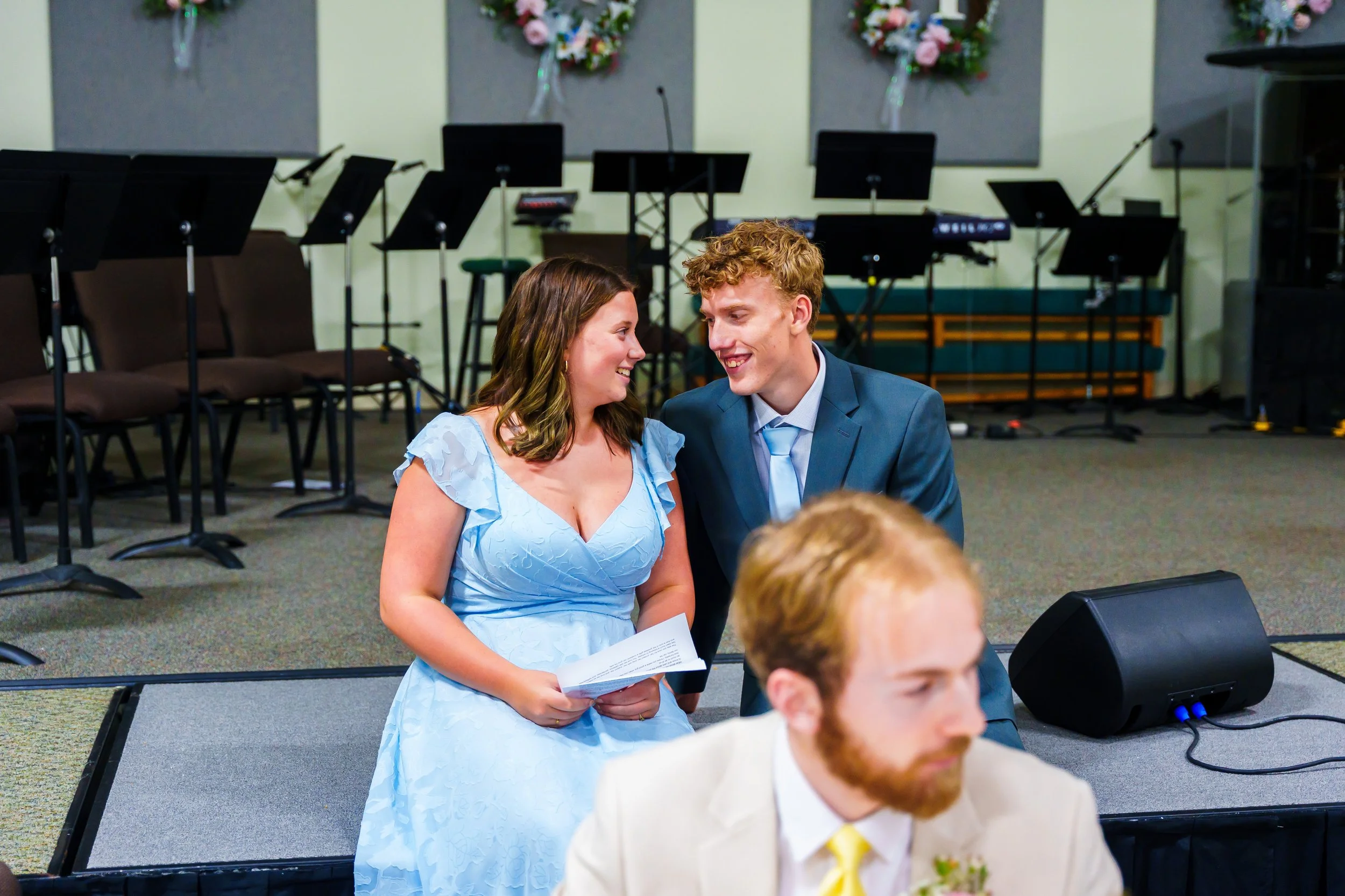 A woman in a light blue dress and a man in a blue suit are smiling and talking to each other at a wedding or formal event. Other guests and musical equipment are visible in the background.