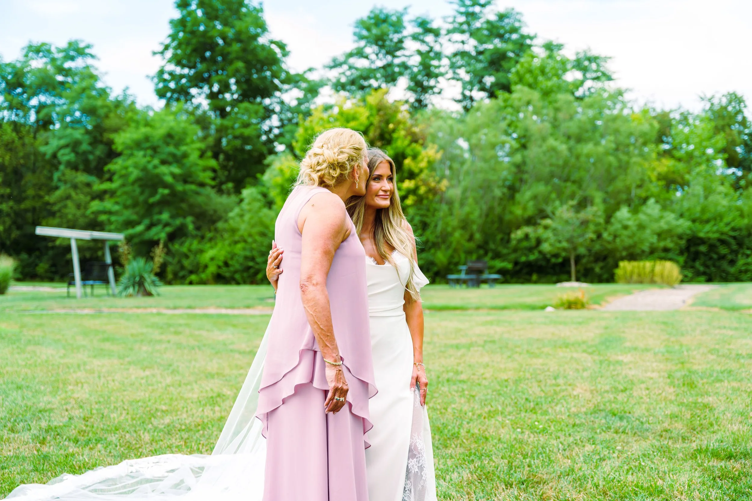 Two women standing on grass in a park, one in a pink dress and the other in a white dress, with green trees in the background.