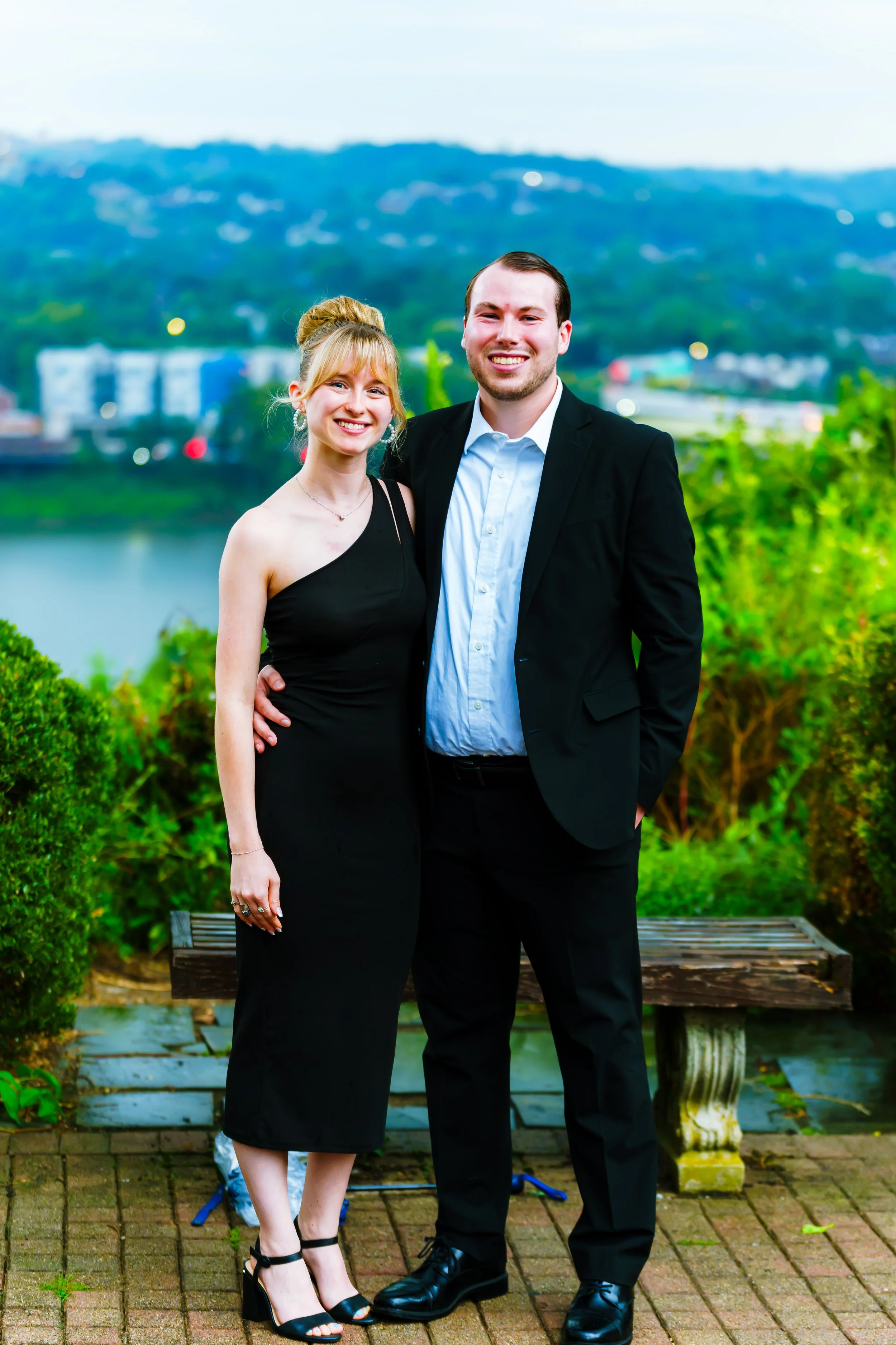 A man and woman dressed in formal attire standing outdoors on a brick path with green bushes and a bench behind them, with a body of water and hills in the background, smiling at the camera.