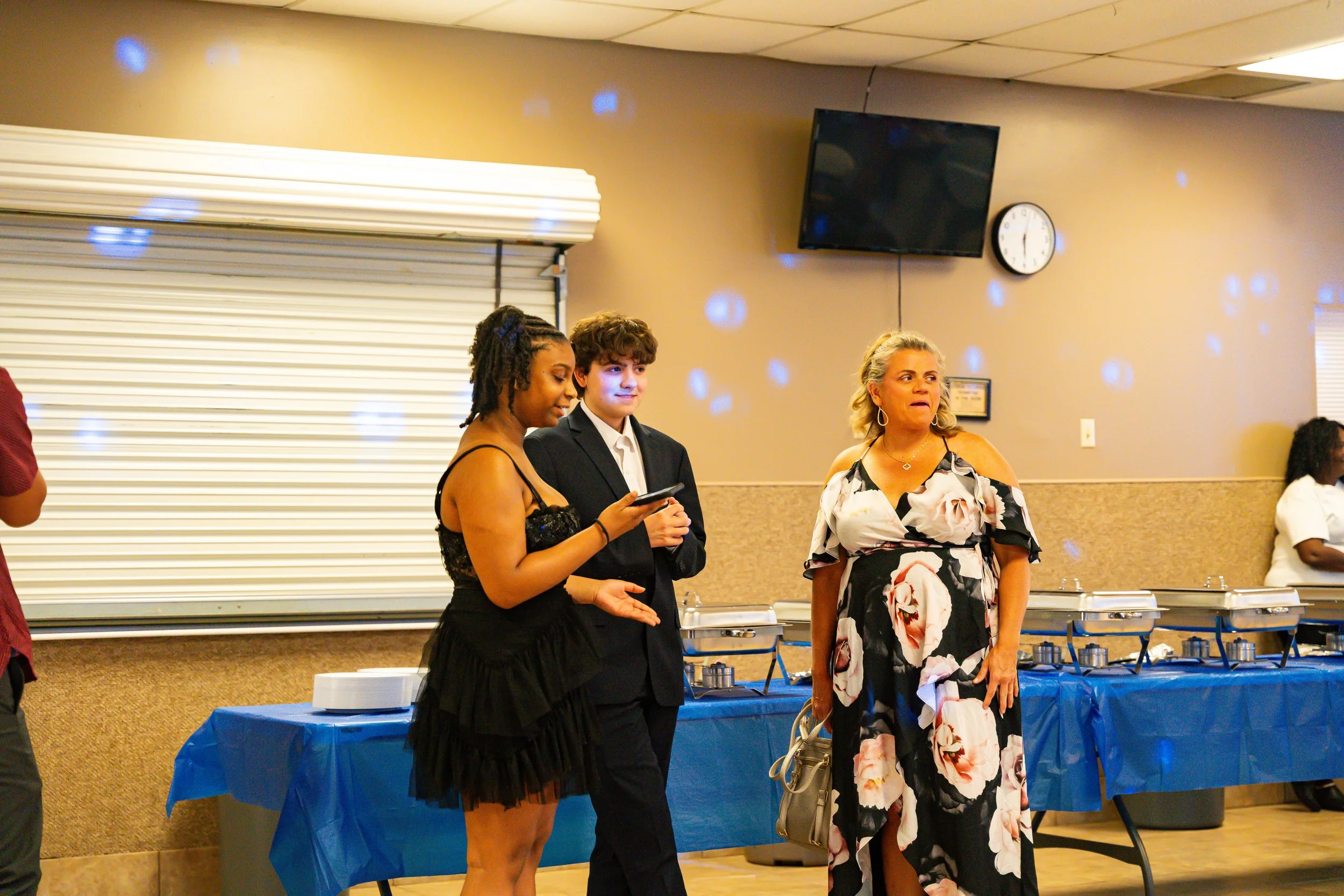 People standing behind a table with trays in a banquet hall, with blue tablecloths and a wall with a clock and a TV in the background.