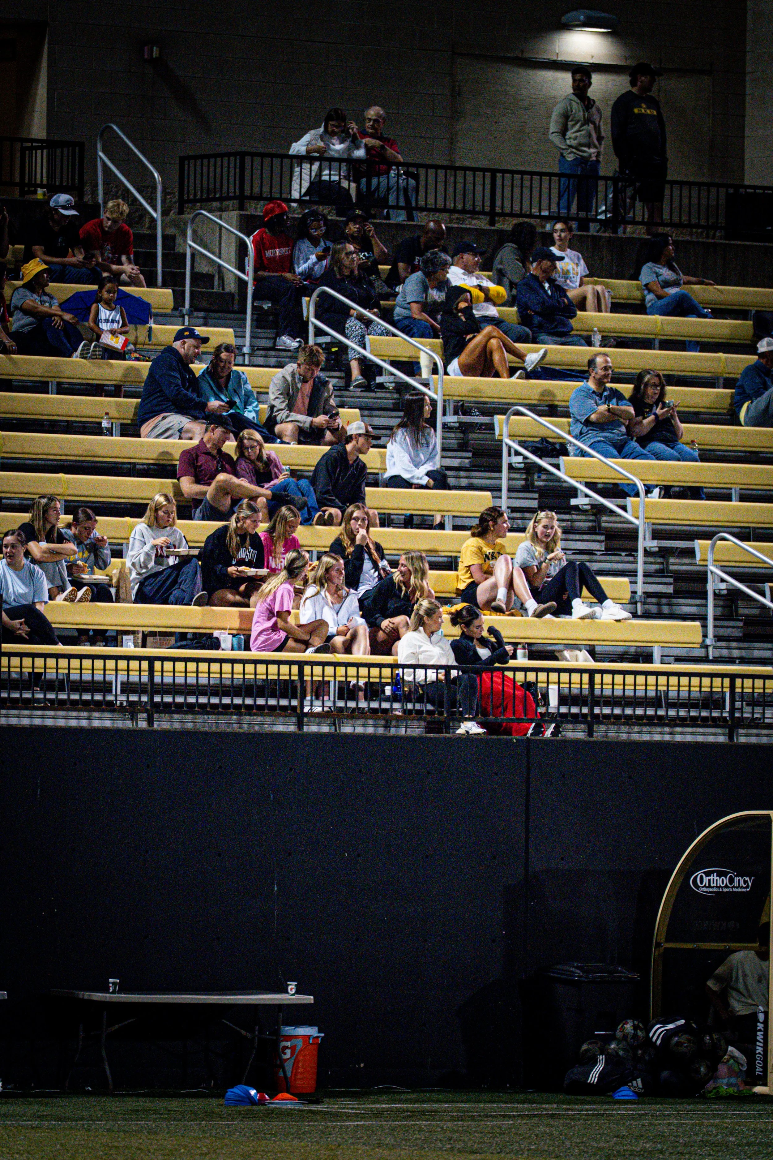 Spectators seated in stadium bleachers watching a sports event, with some using their phones and others talking or eating.
