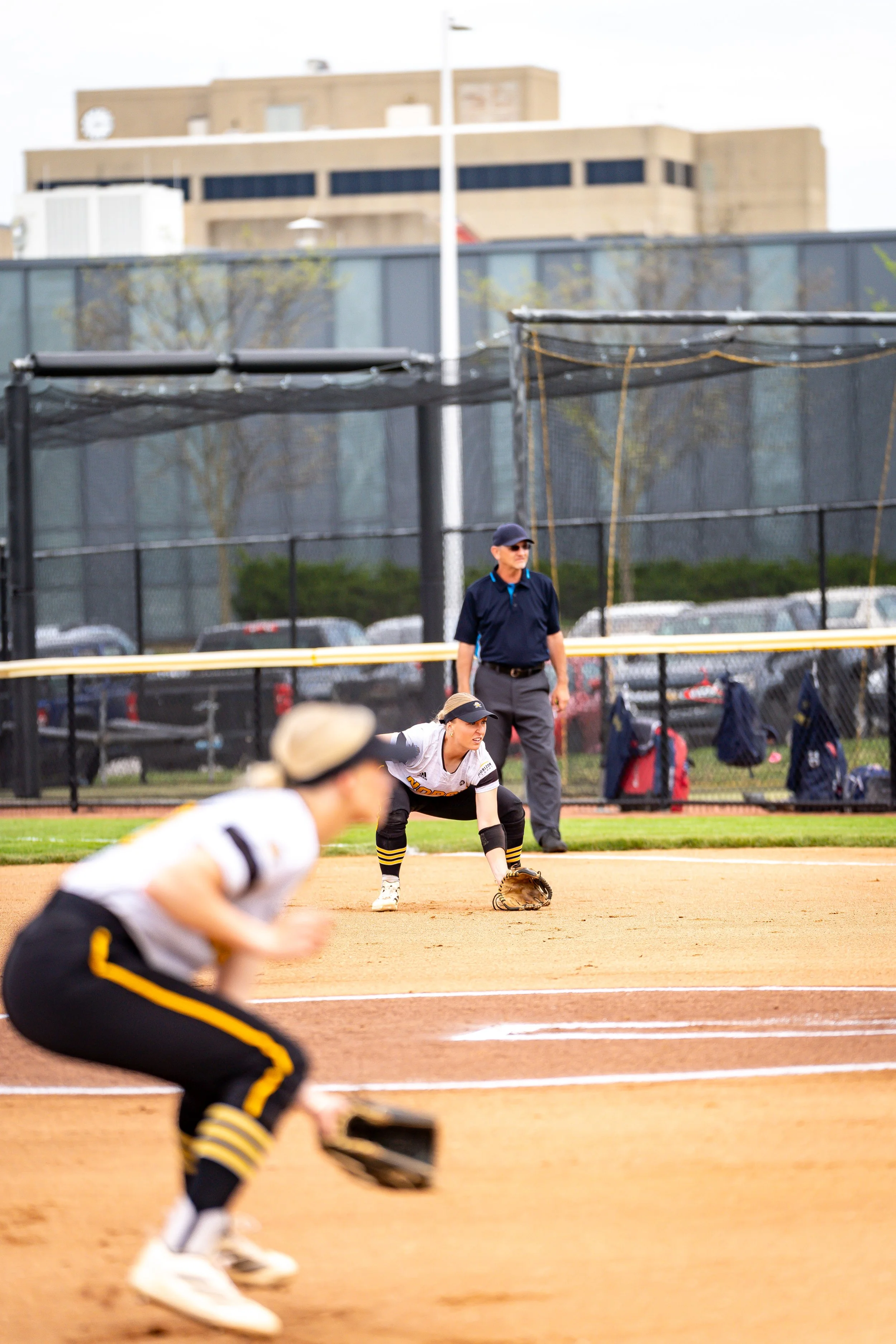 A softball player in a white jersey prepares to field the ball, while other players and a coach watch from the background on the field.