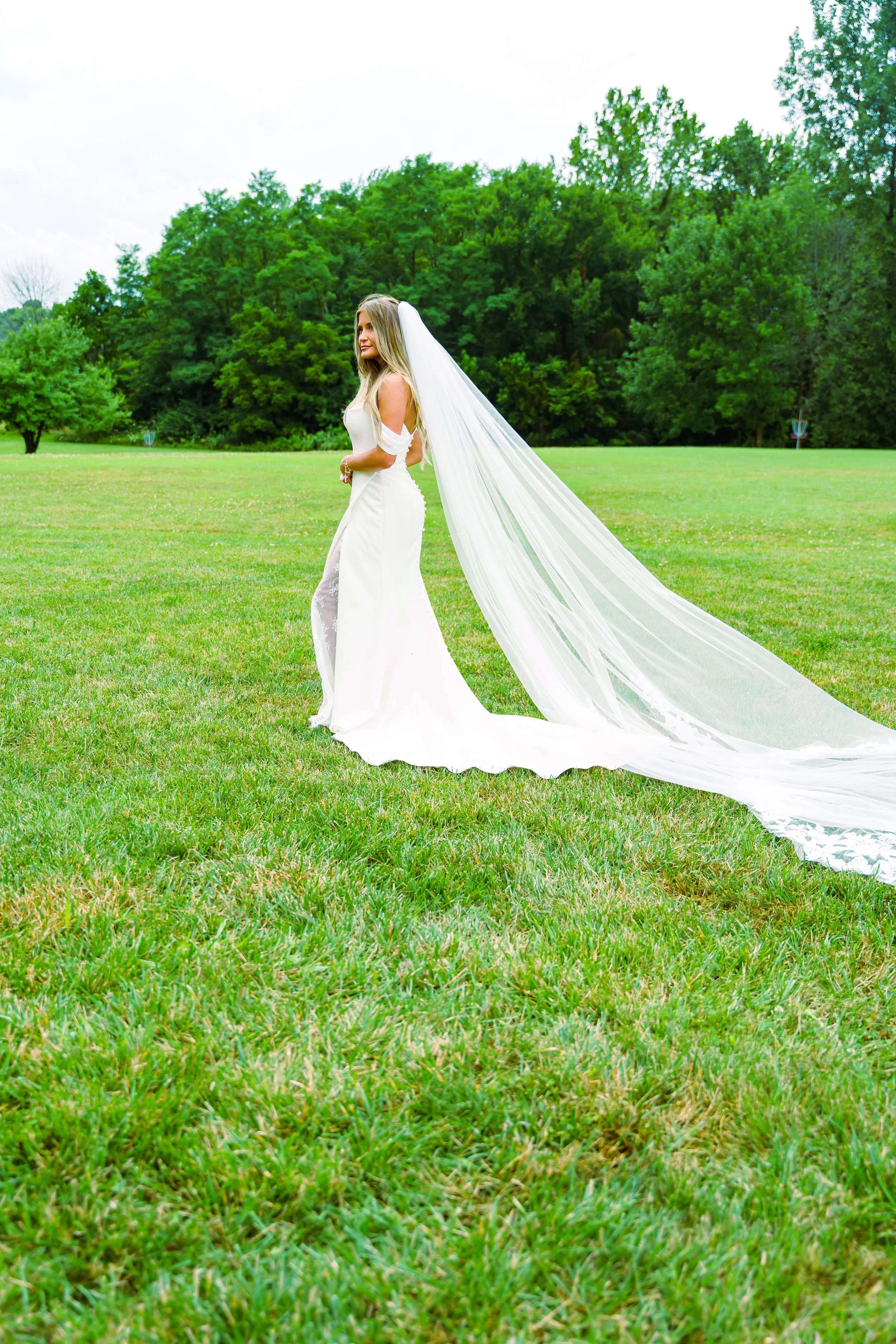 A bride in a white wedding gown with a long train and veil standing on a grassy field with green trees in the background.