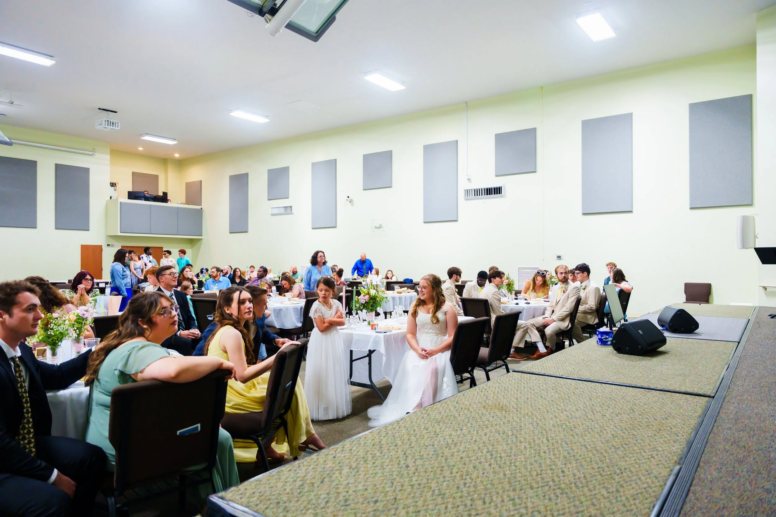 Guests seated at round tables in a large, well-lit banquet hall attending a formal event or wedding reception; a bride in a white dress is standing near the center.