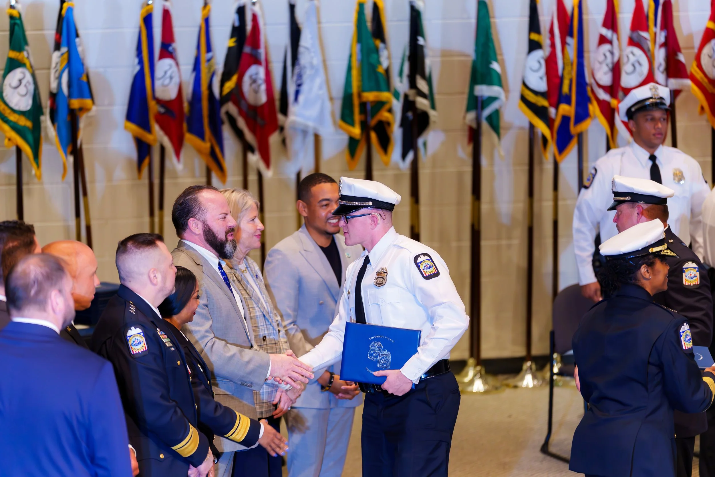 People in police uniforms and formal attire at an awards ceremony, with flags in the background.