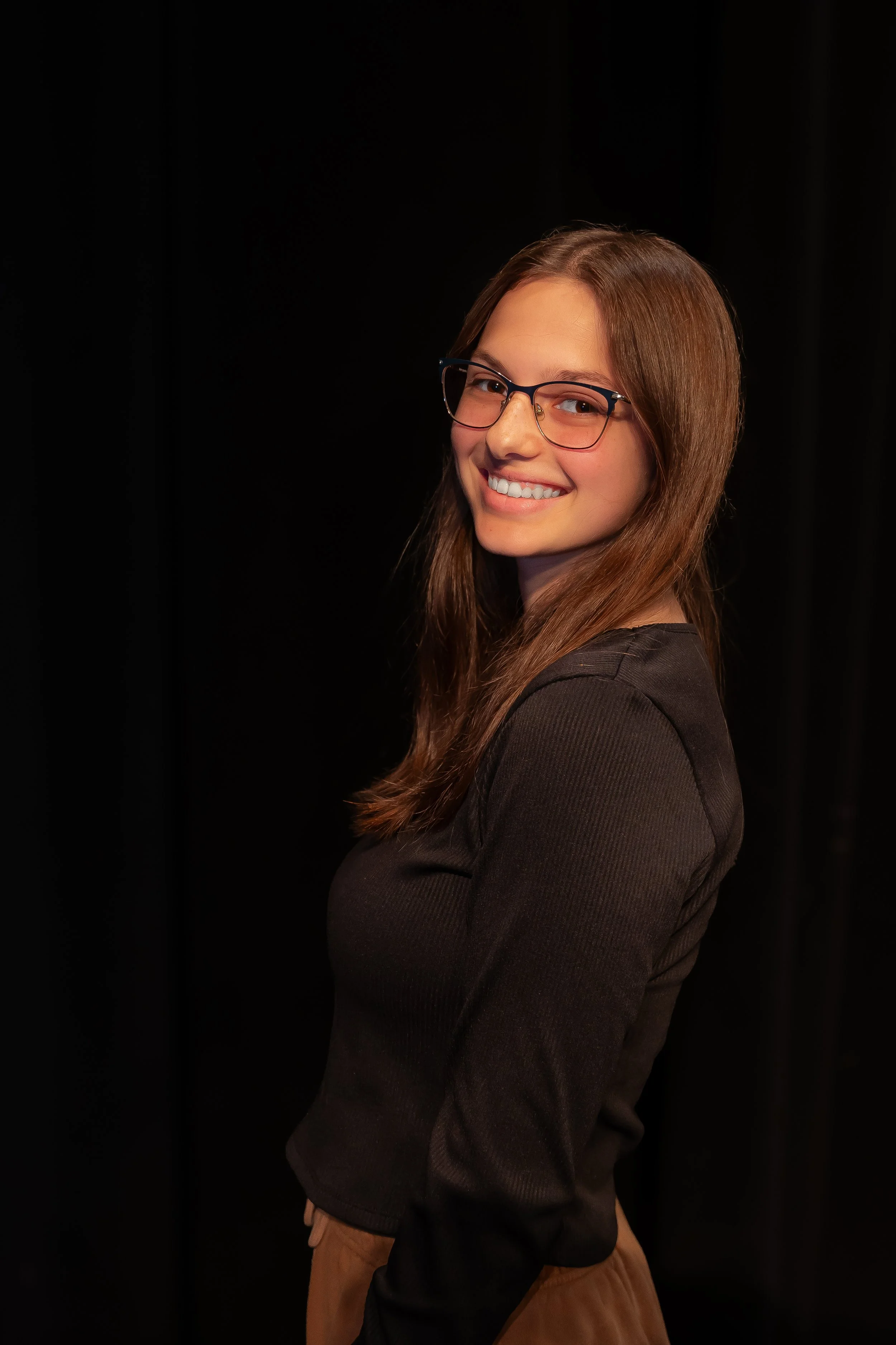A woman with long brown hair, glasses, and a dark top, smiling and looking at the camera against a black background.