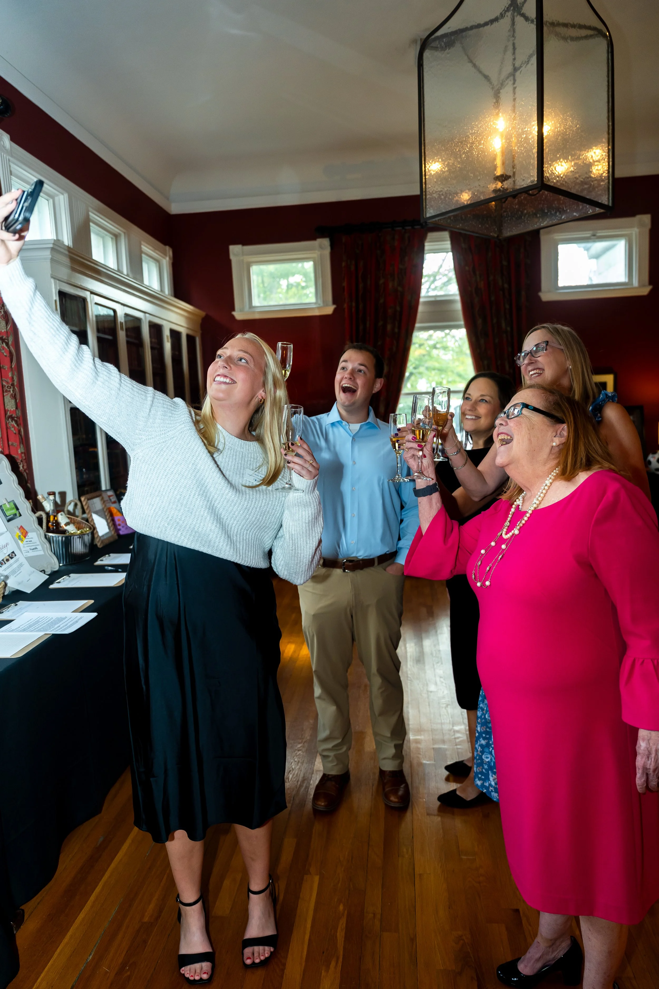 A group of six people, five women and one man, are taking a selfie together at an indoor gathering. They are smiling and holding glasses of champagne, dressed in elegant attire, in a room with red walls, windows, and a large chandelier.