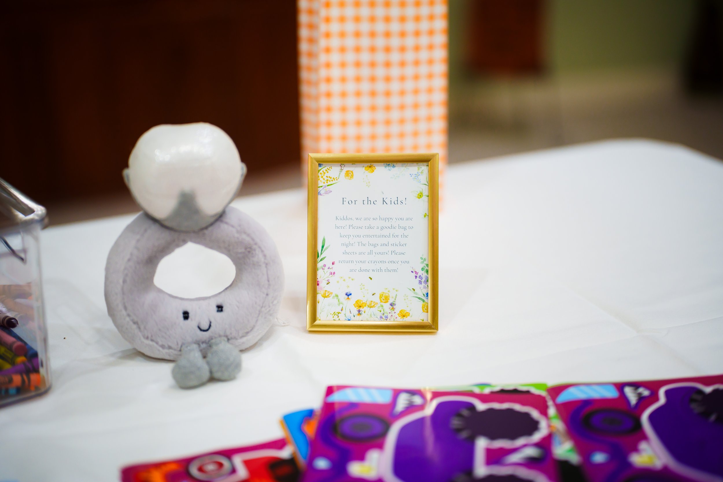 Decorative sign titled "For the Kids!" with floral border, a whimsical figurine of a smiling character with a round white head, glasses, and a purple body, small gray shoes, on a white table among colorful candy wrappers and a container of colored pe
