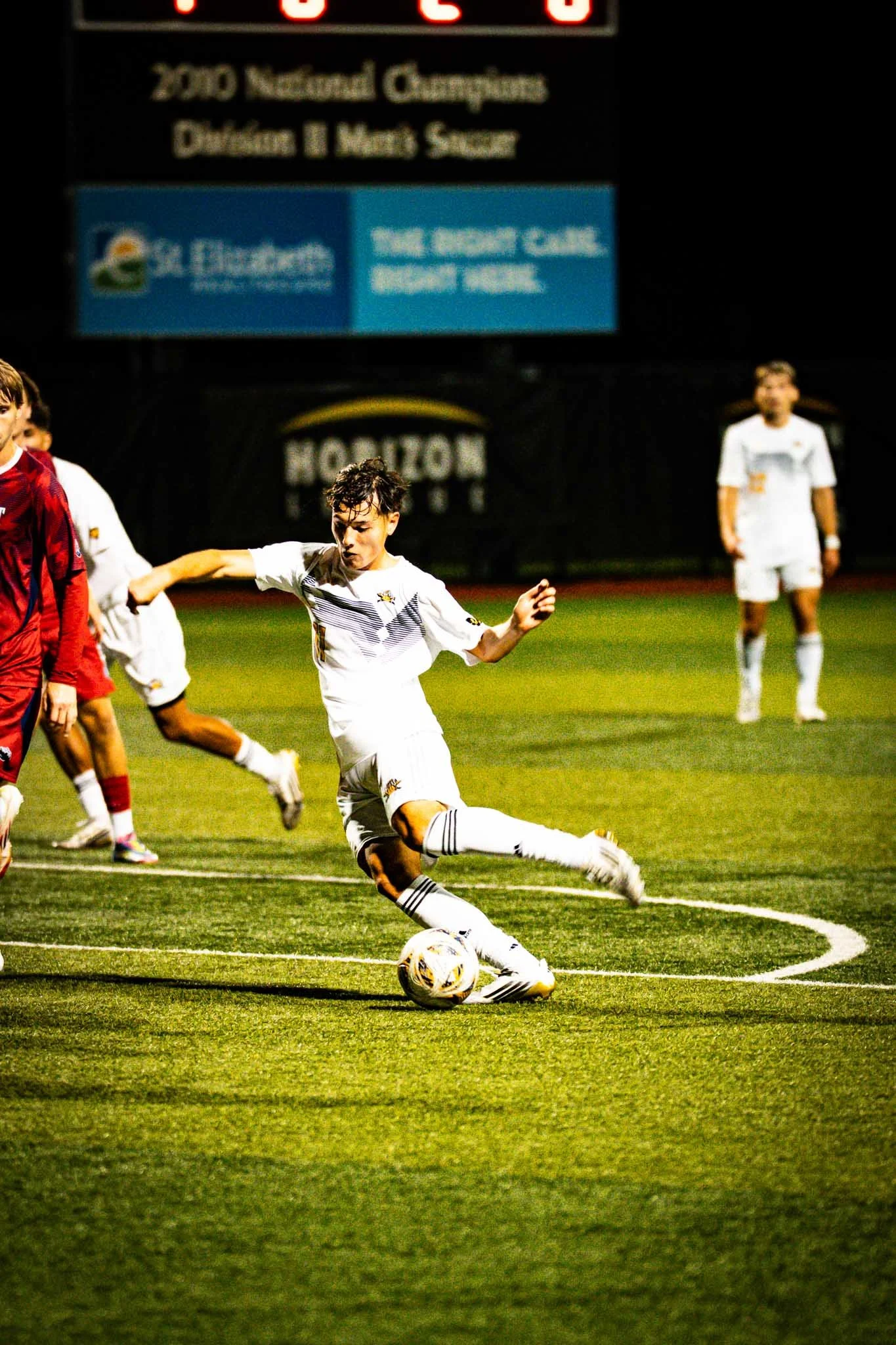 A young soccer player in a white uniform is kicking a soccer ball on a field during a night game, with other players in the background.